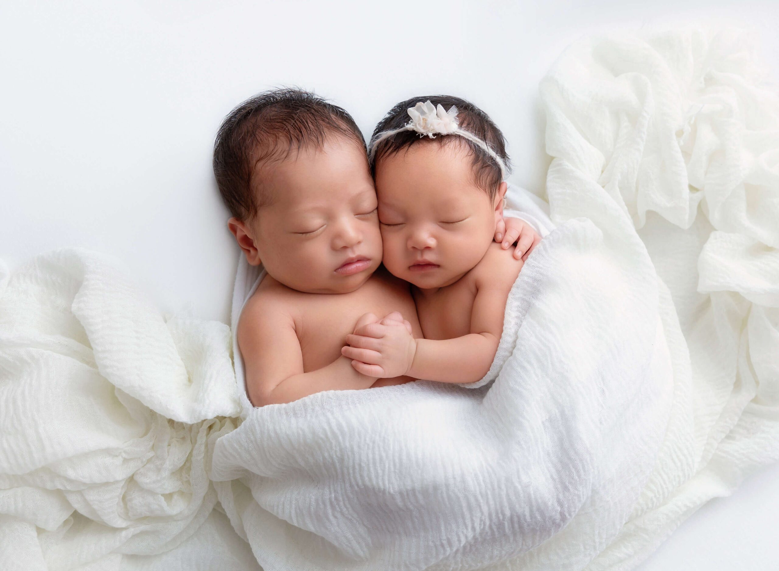 Newborn twins of boy and the girl laying on a white blanket holding hands 