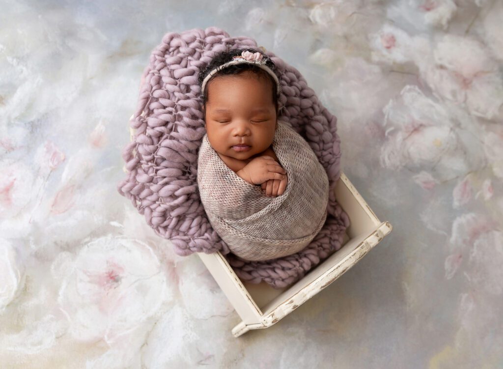 Newborn baby girl asleep in miniature crib with purple floral background