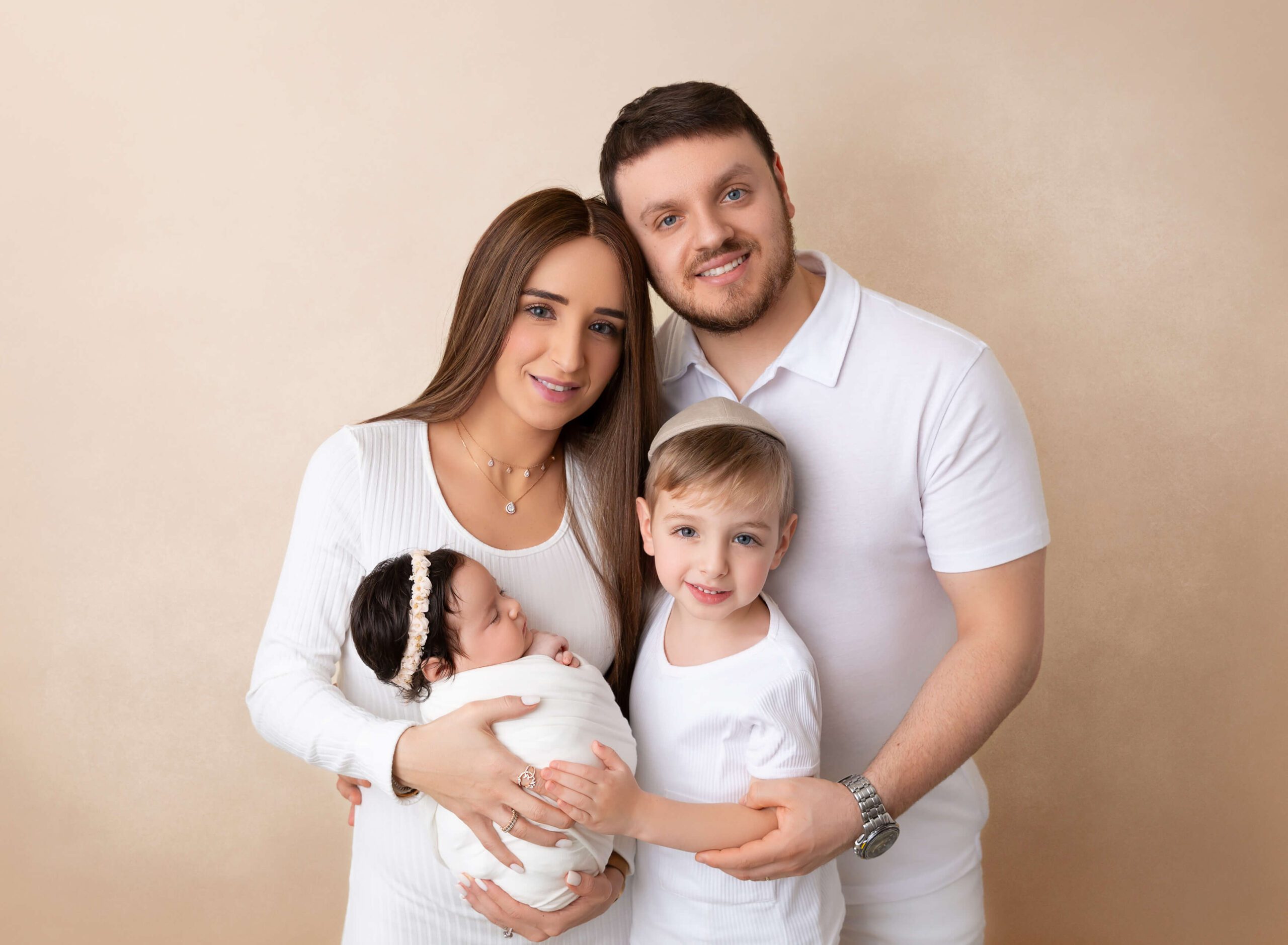 A happy mom and dad stand in a studio holding their newborn baby and toddler son