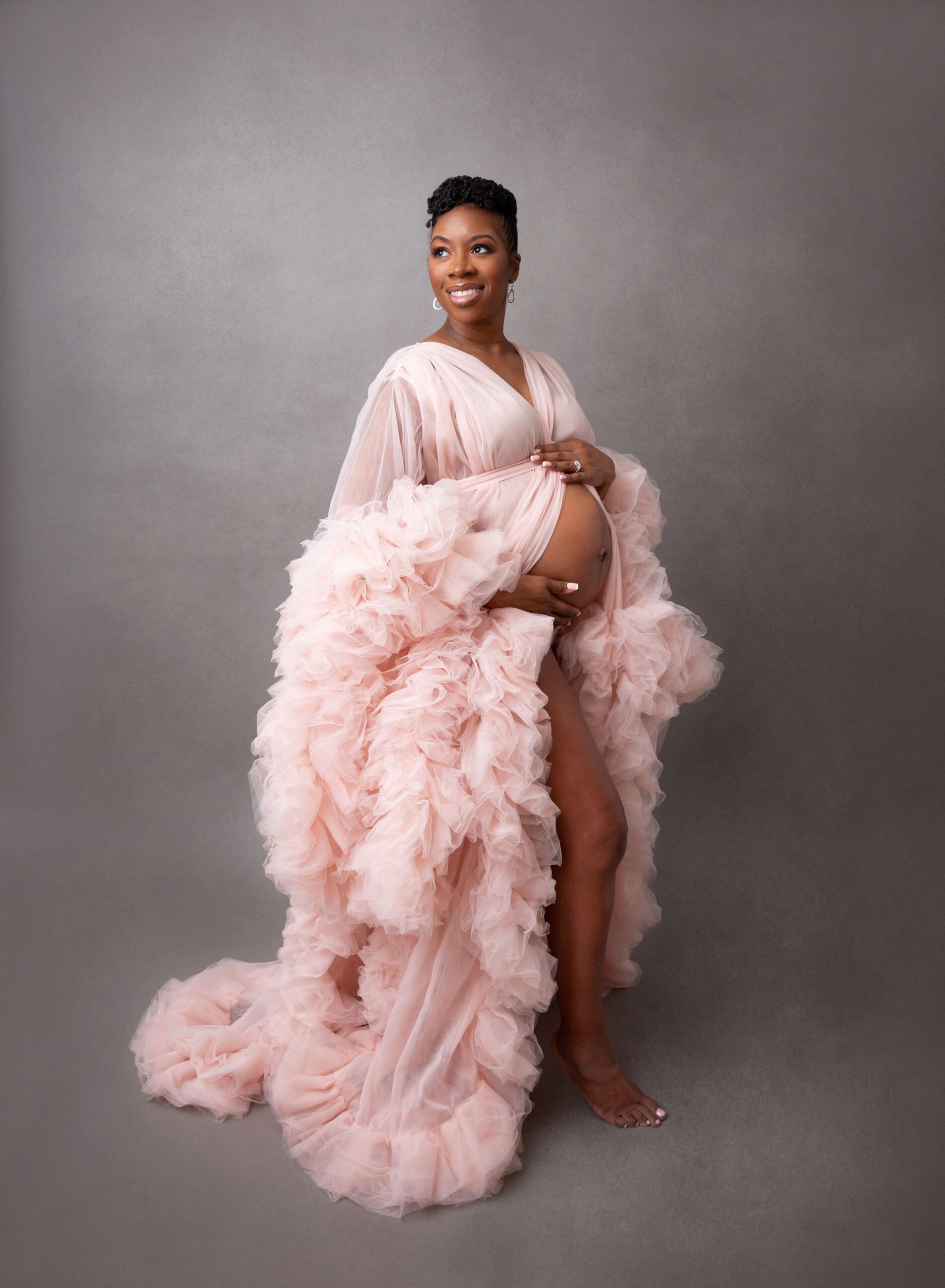 A happy mother to be smiles over her shoulder while holding her bump in a pink tule gown in a studio