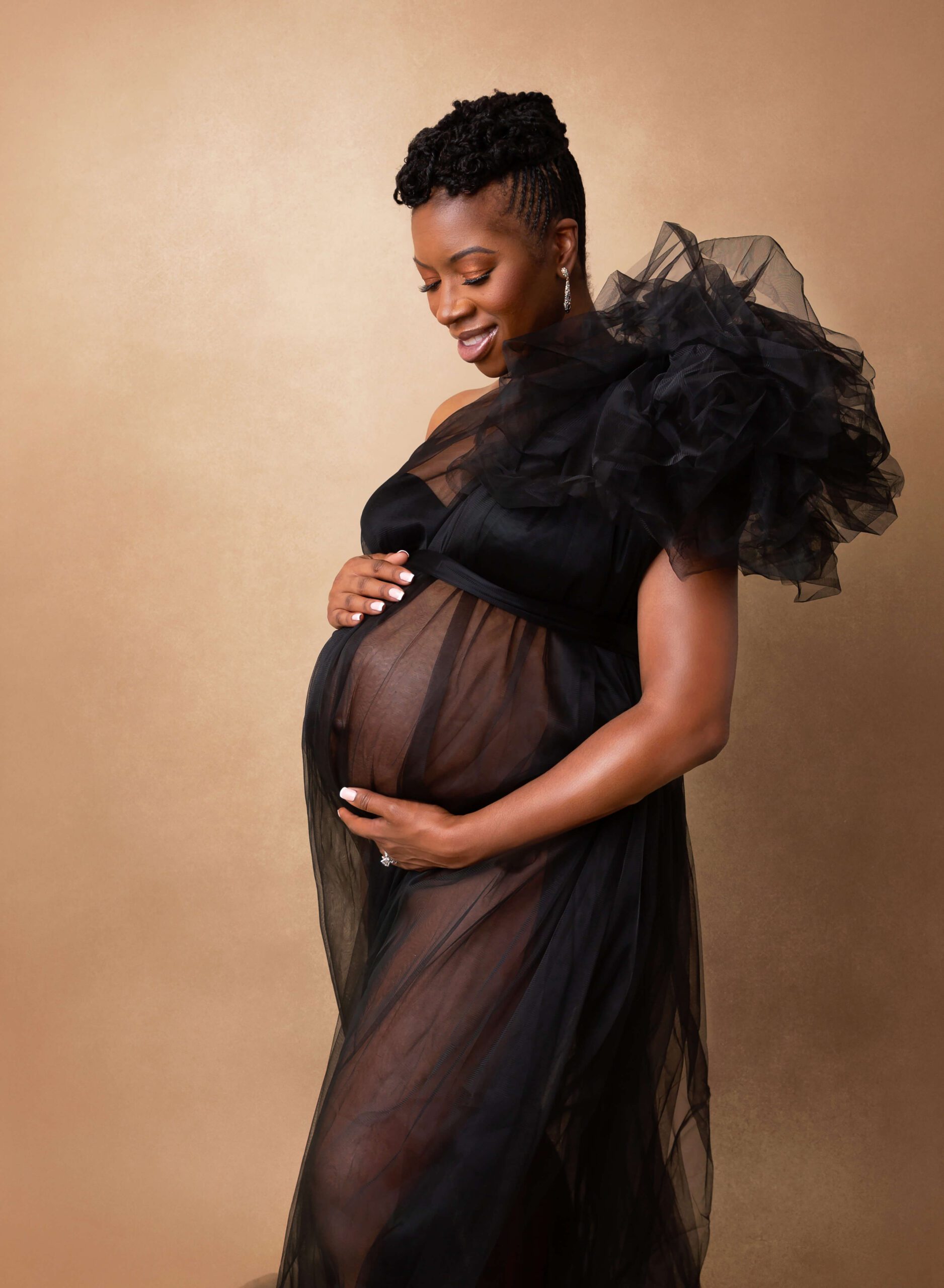 A smiling mother to be gazes down to her bump while holding it in a studio in a black tule gown after meeting Brooklyn midwives