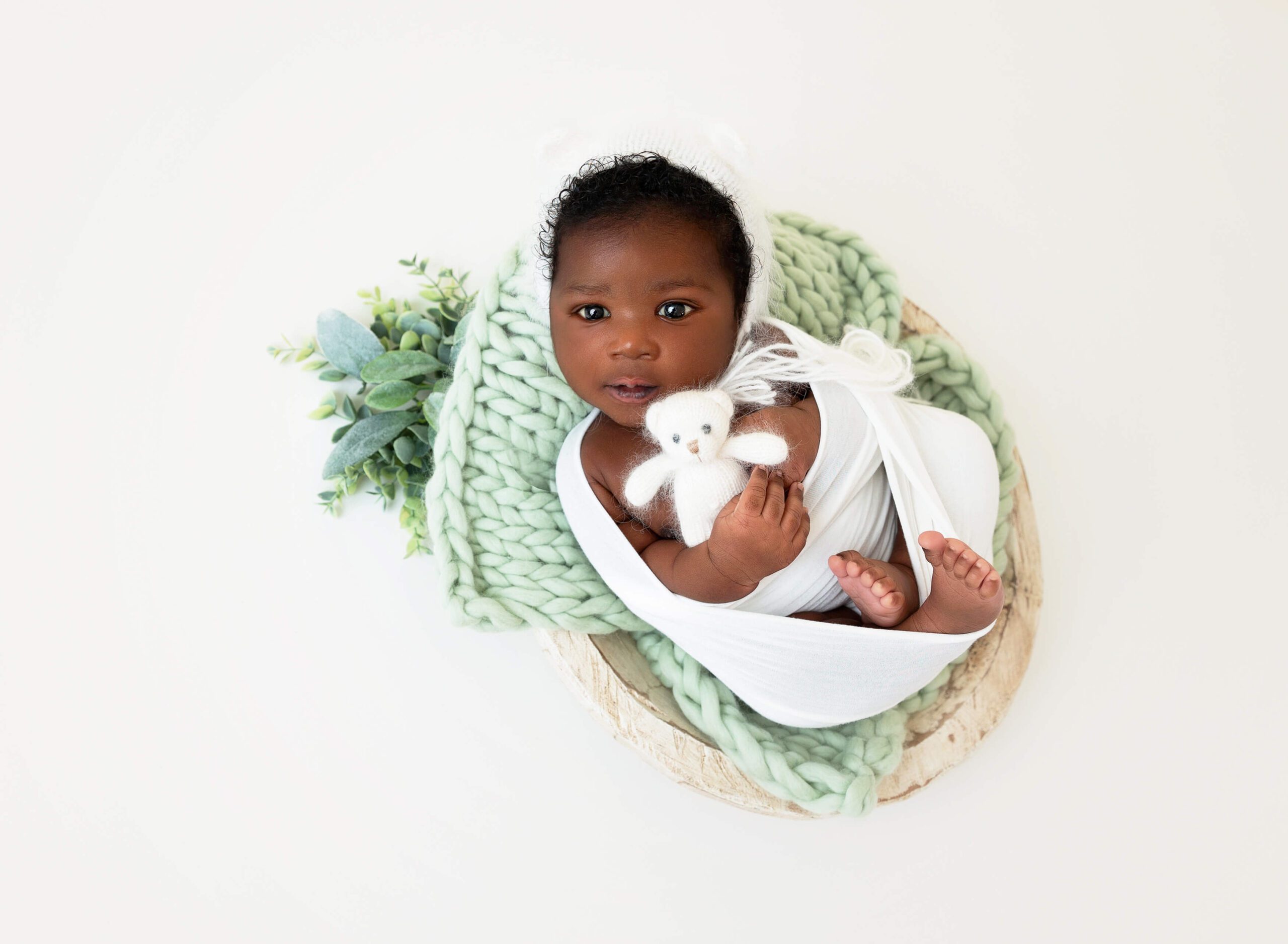 A newborn baby lays in a blanket with eyes open on green blankets in a white swaddle holding a knit bear in a studio