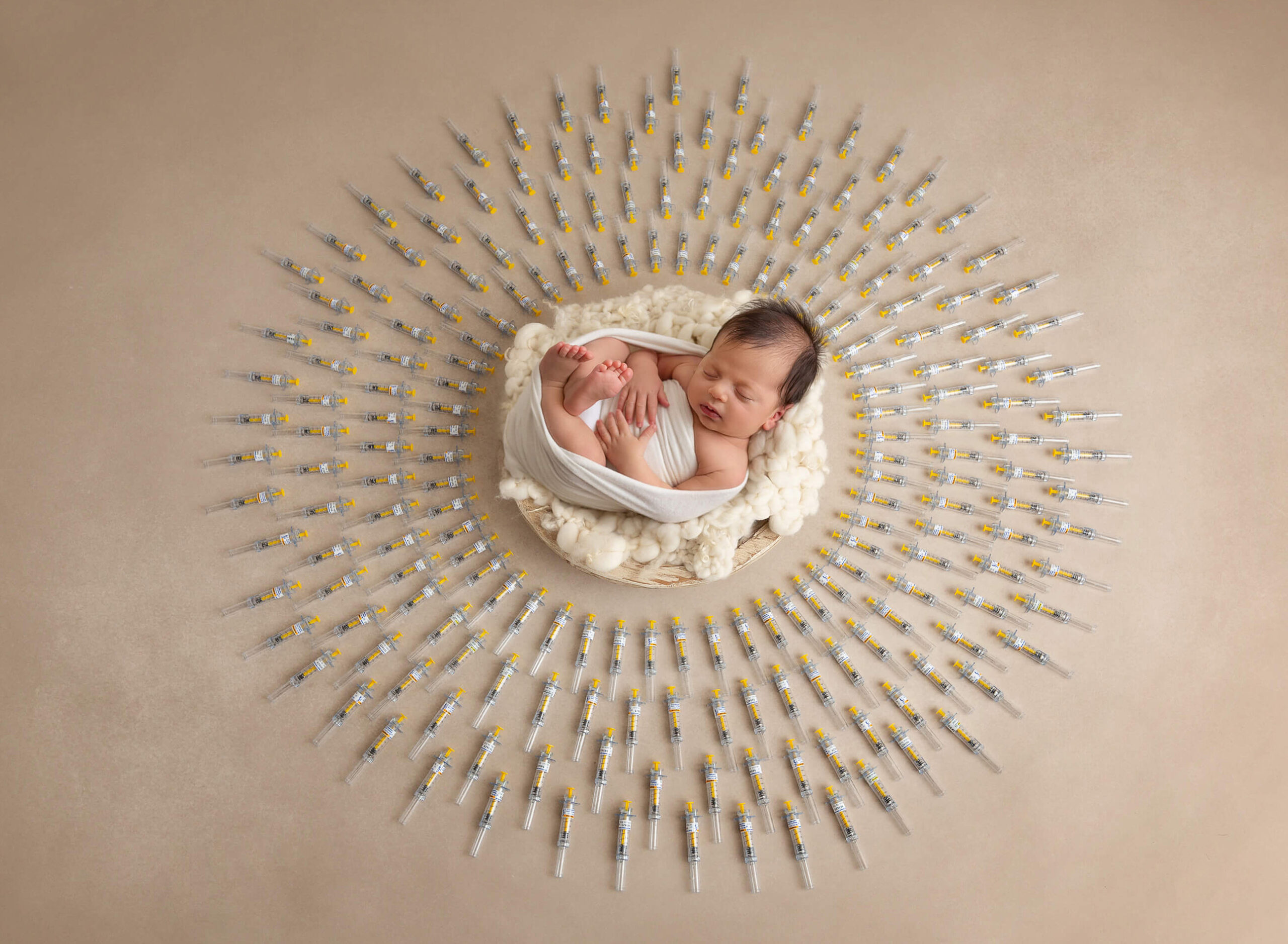 A newborn baby peacefully swaddled in a white wrap, lying on a soft cream-colored blanket. The baby is surrounded by a radiant, circular arrangement of syringes placed evenly, creating a symbolic and artistic display.