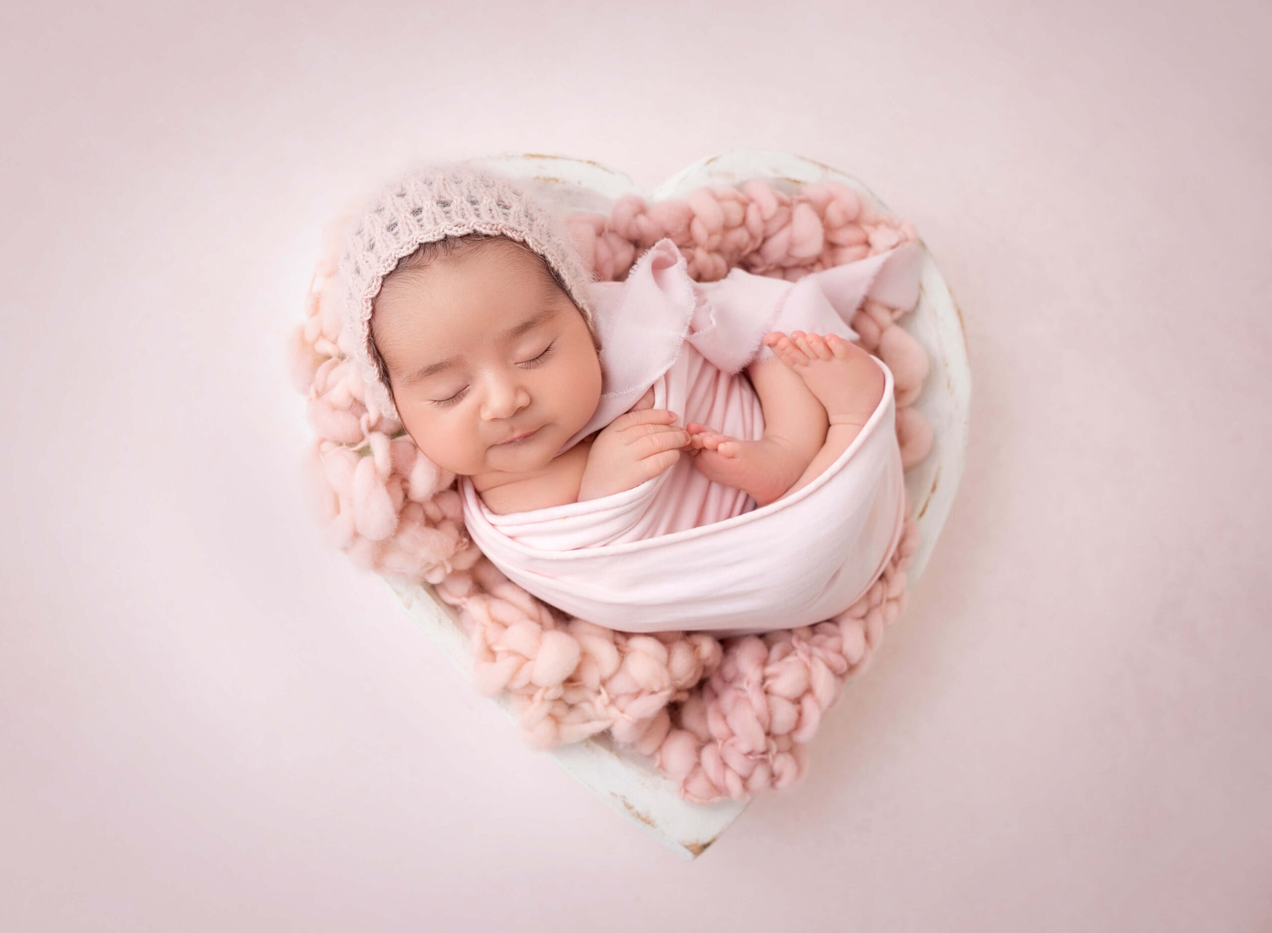 A newborn baby lays in a winy wooden heart shaped bowl  in a pink swaddle thanks to fertility clinics in new york
