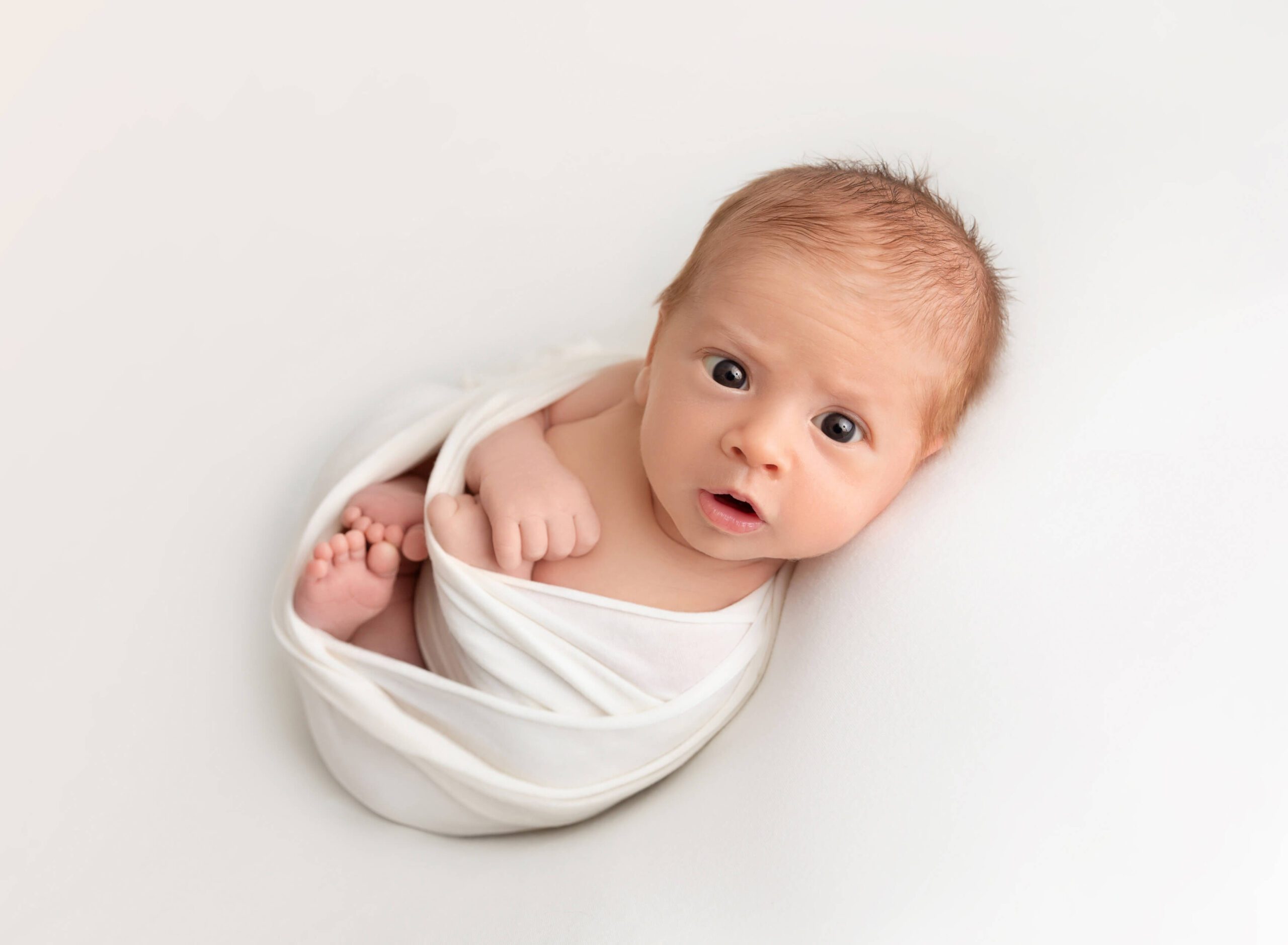 An awake newborn baby lays on a white bed in a white swaddle with eyes wide open after meeting doulas in new york