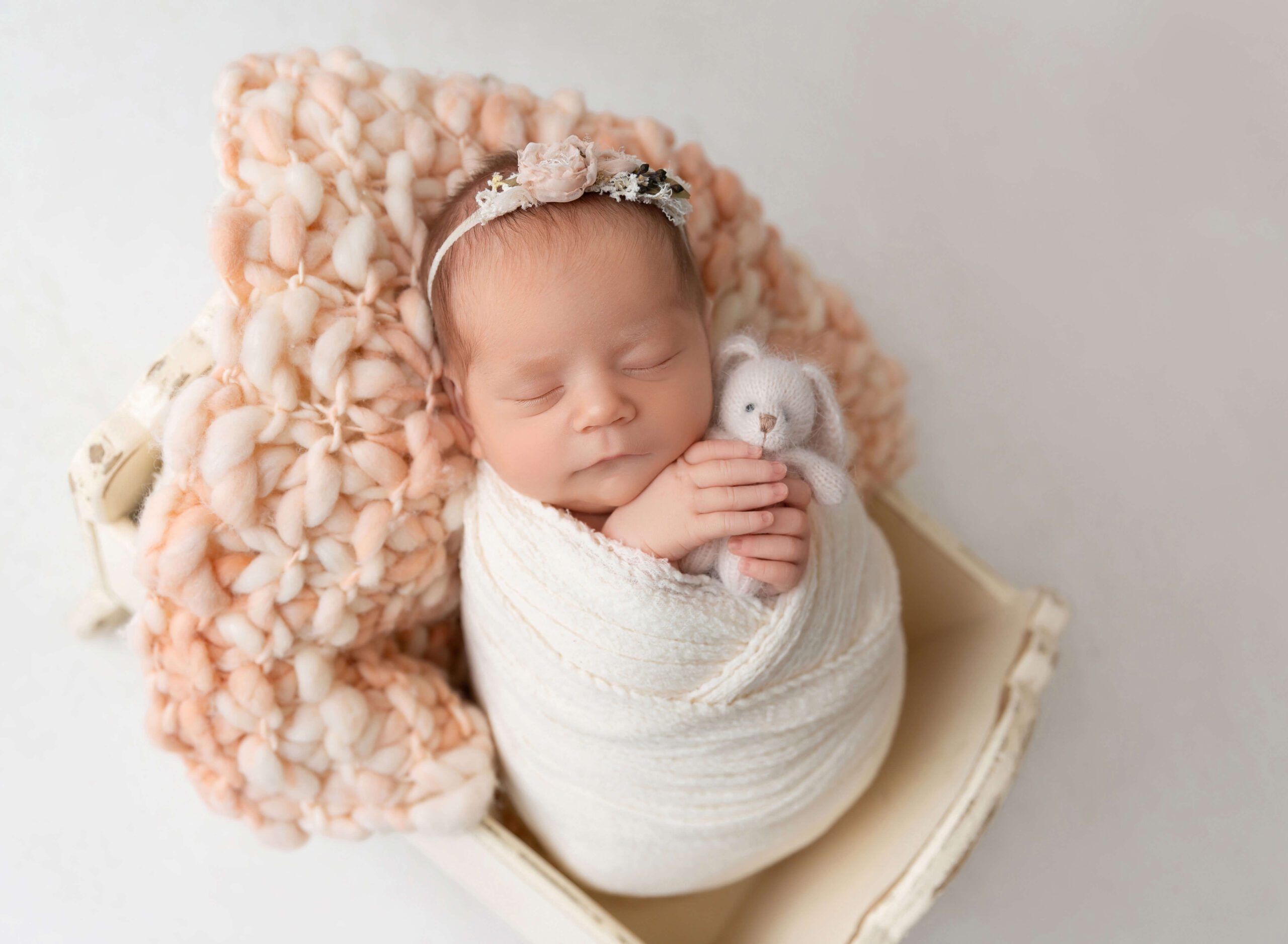 A newborn baby girl sleeps holding a knit bunny in a tiny wooden crib and white swaddle after using doulas in new york