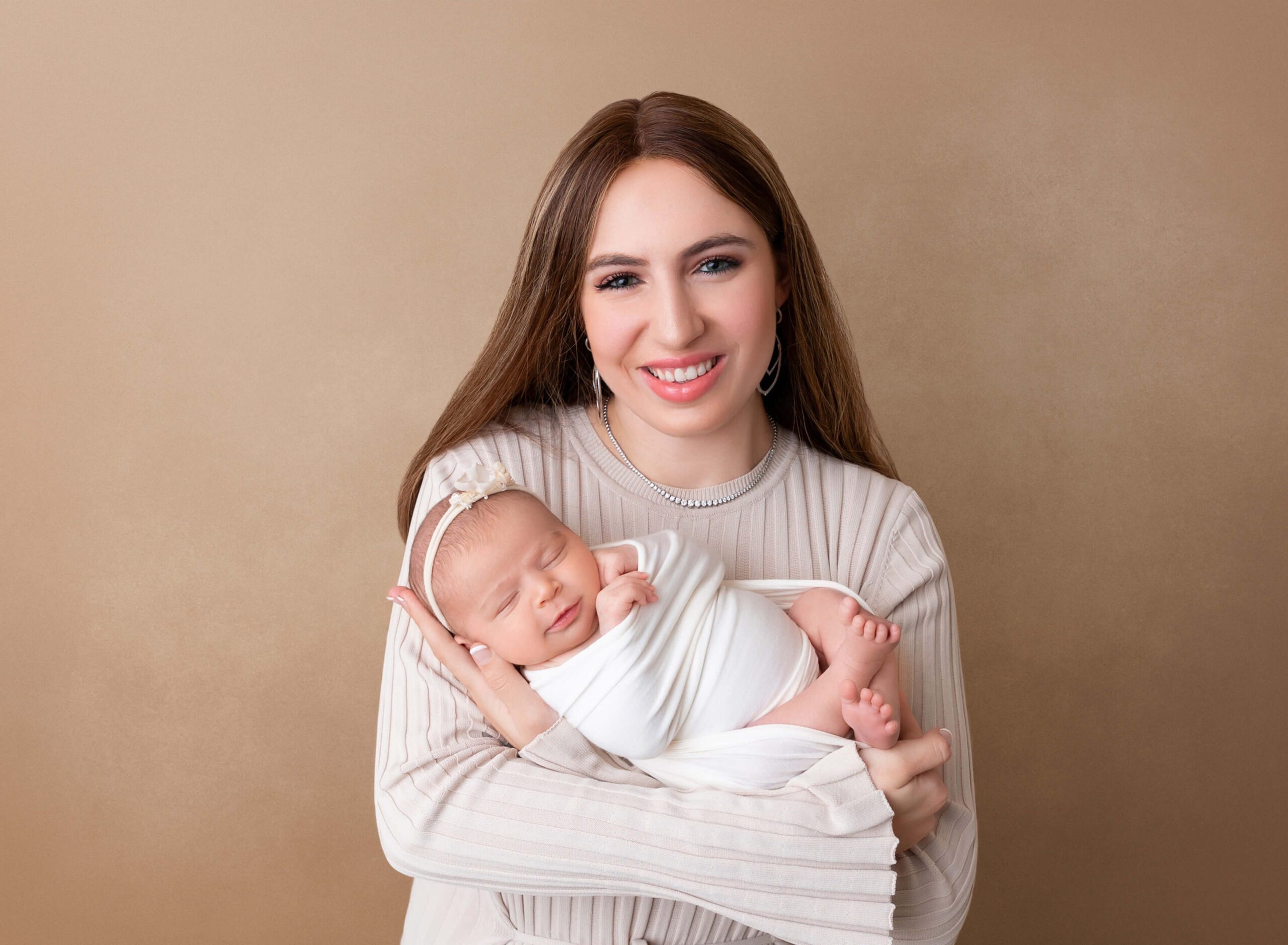 A happy new mom cradles her sleeping newborn baby in her arms in a white swaddle