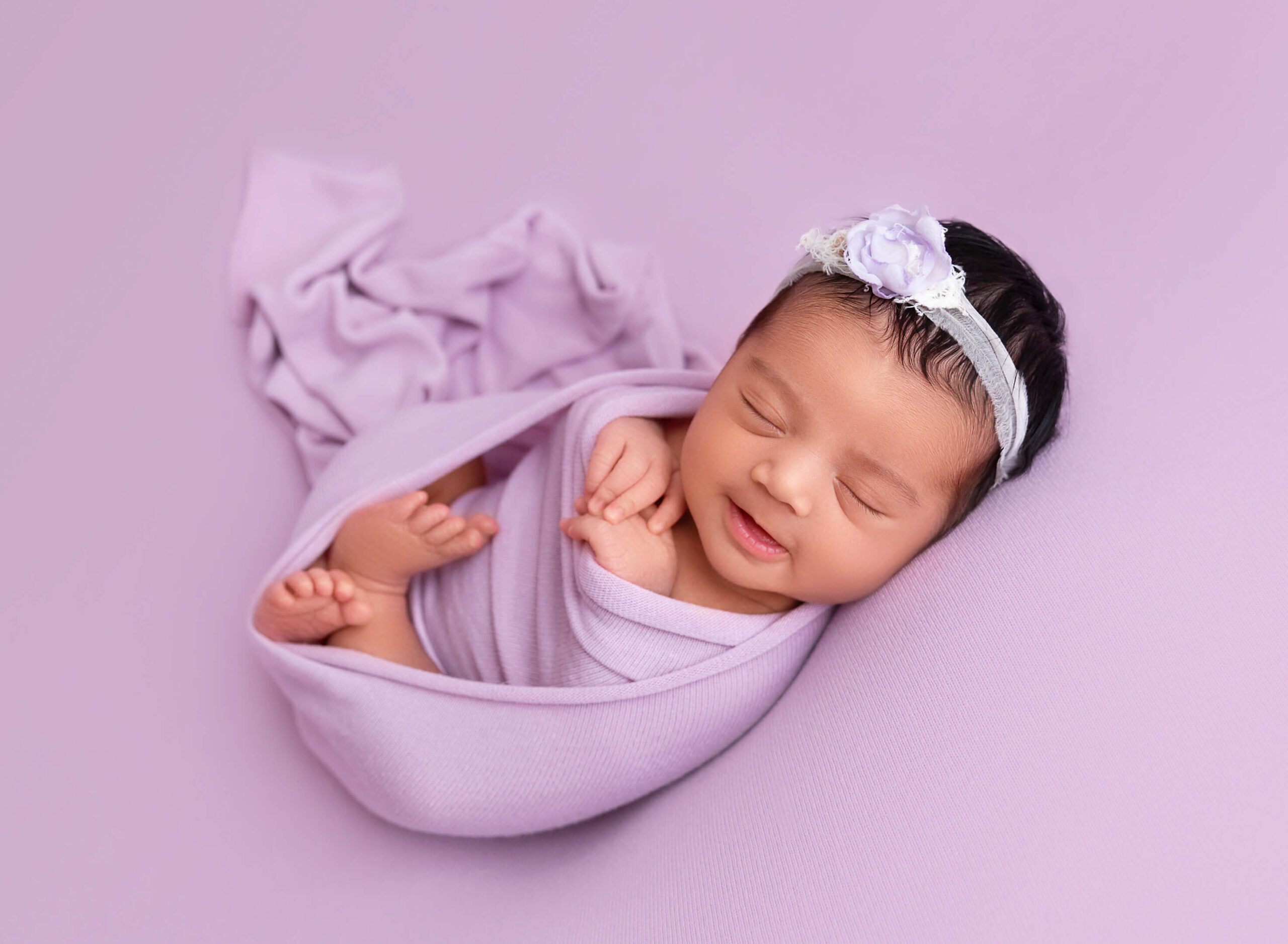 A smiling newborn baby sleeps in a purple swaddle and matching floral headband in a studio after using midwives in new york