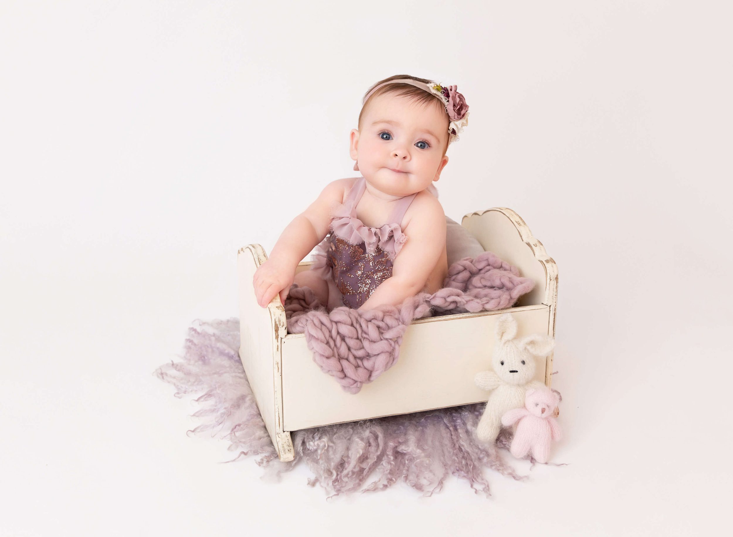 A baby girl in a purple dress sits in a tiny wooden crib with knit bunnies