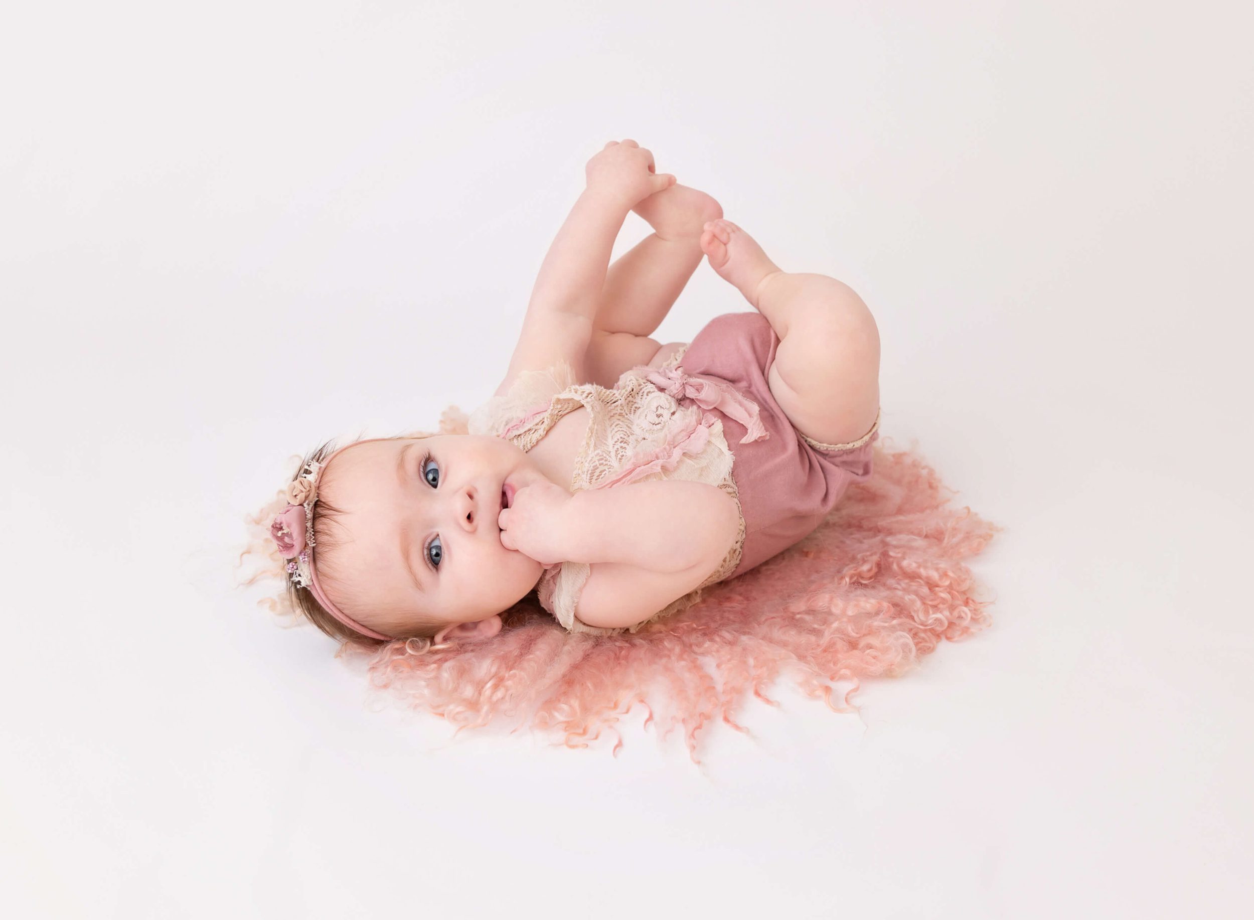 A baby plays with her feet in a pink onesie while on her back on the floor of a studio