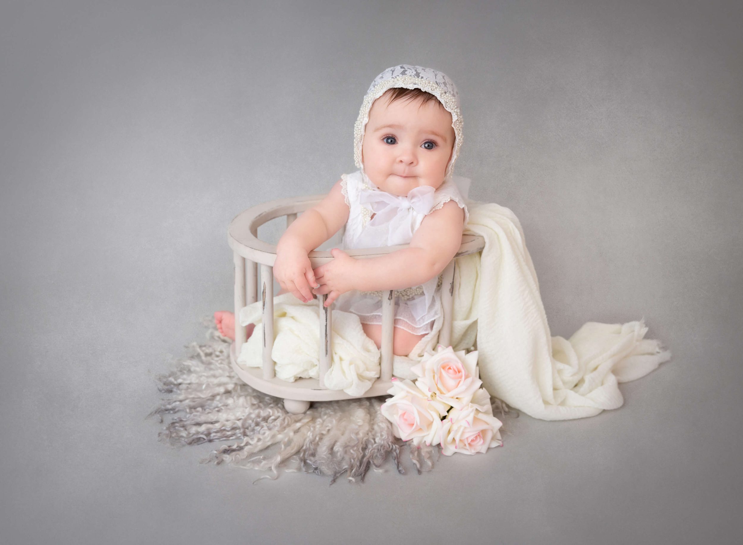 A baby sits in a tiny wooden crib in a lace bonnet and white dress after meeting nannies in New York