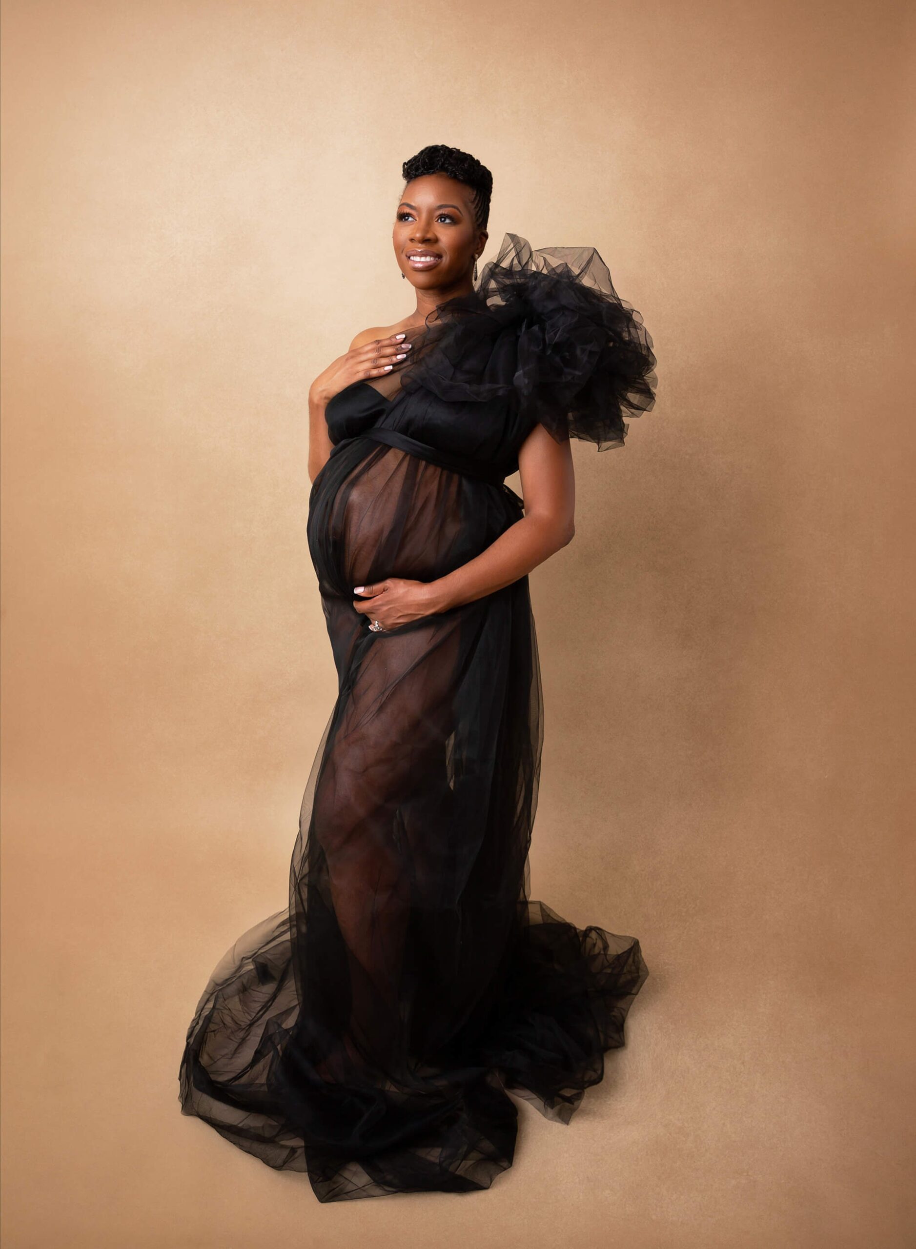 A happy mom to be stands in a studio in a black tule maternity gown