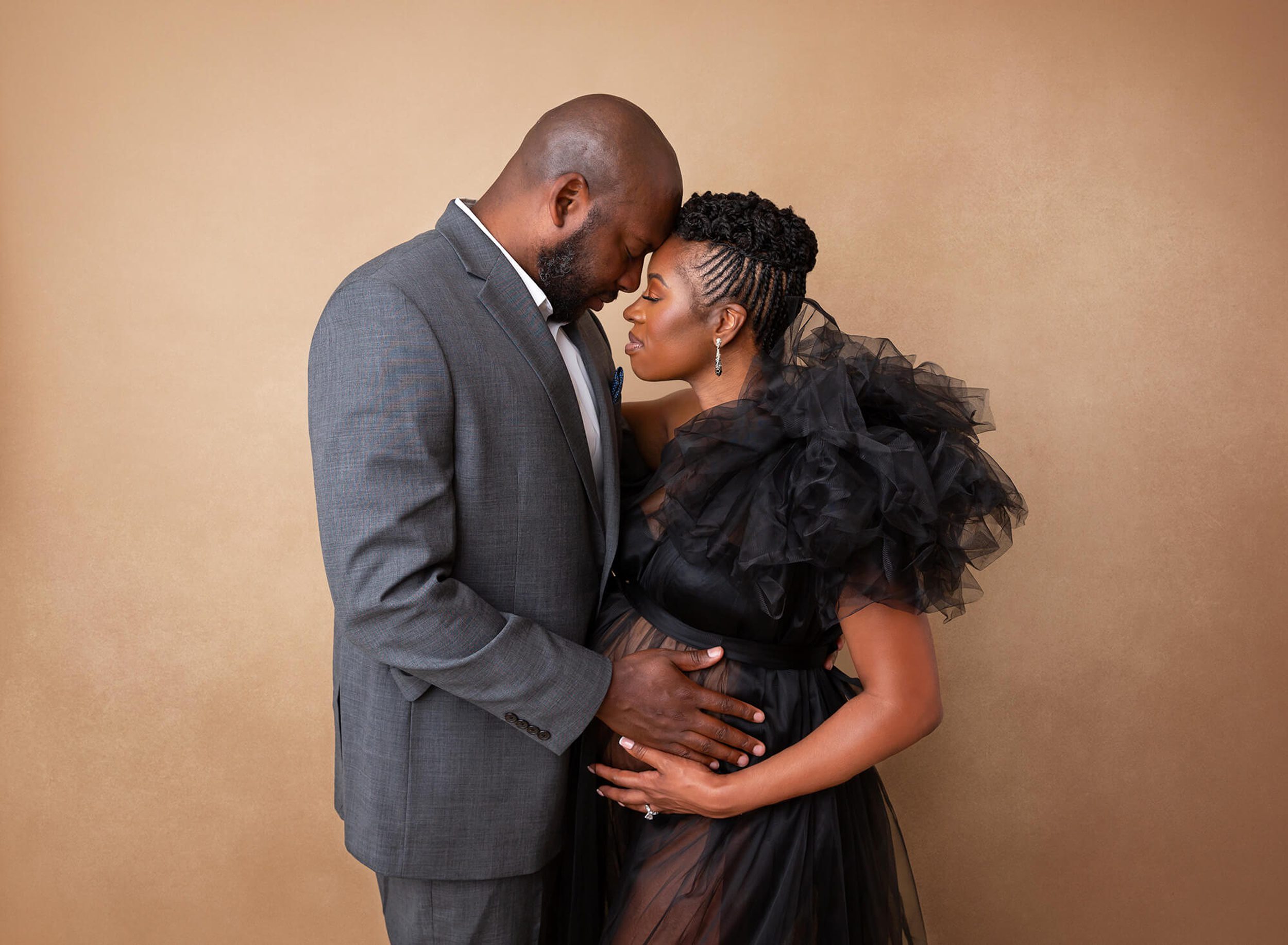 An expecting couple with hands on the bump touch foreheads while standing in a studio in a black tule dress and grey suit after meeting a postpartum doula in new york