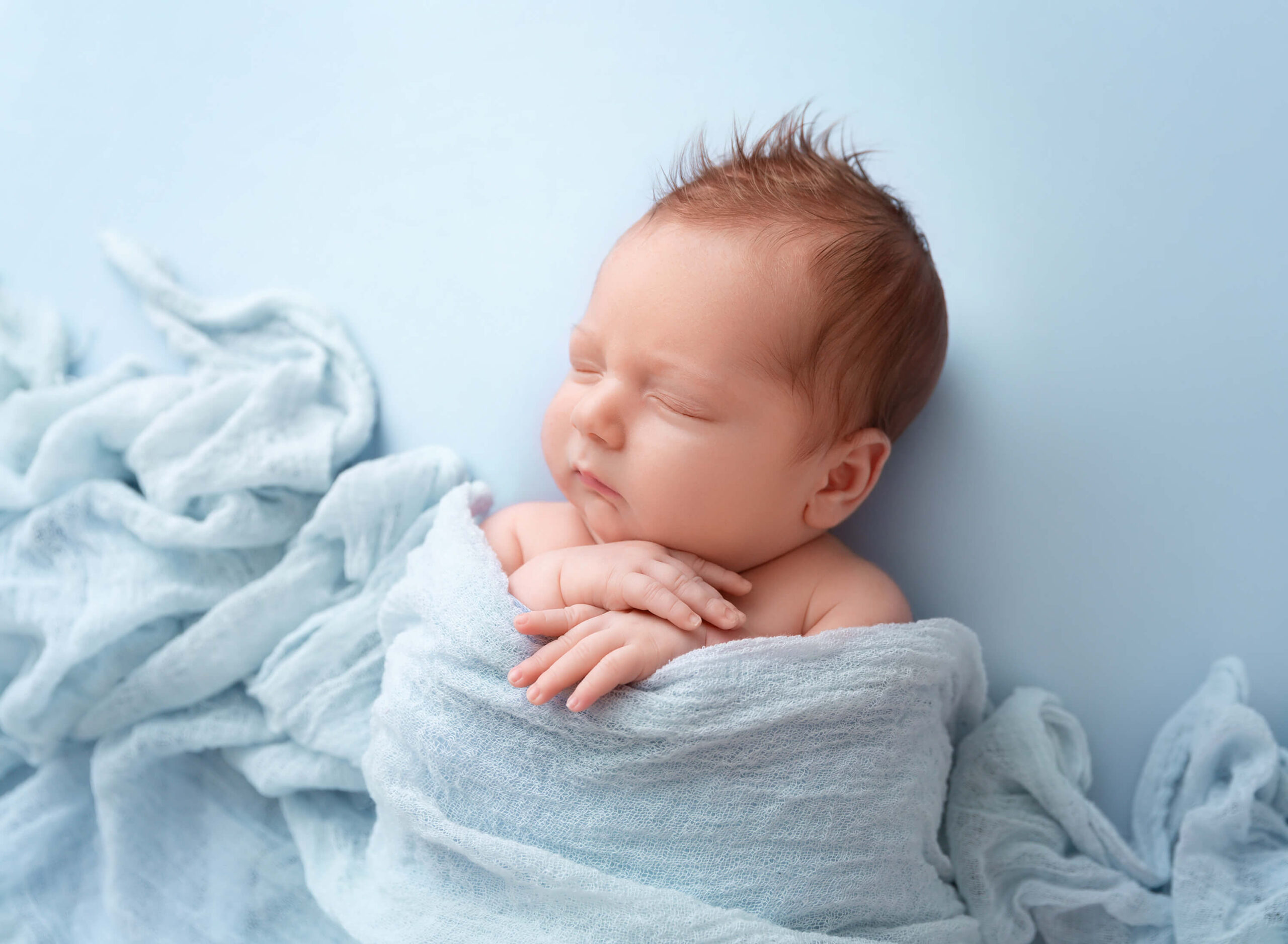 A red hair newborn baby sleeps under a blue blanket with hands crossed