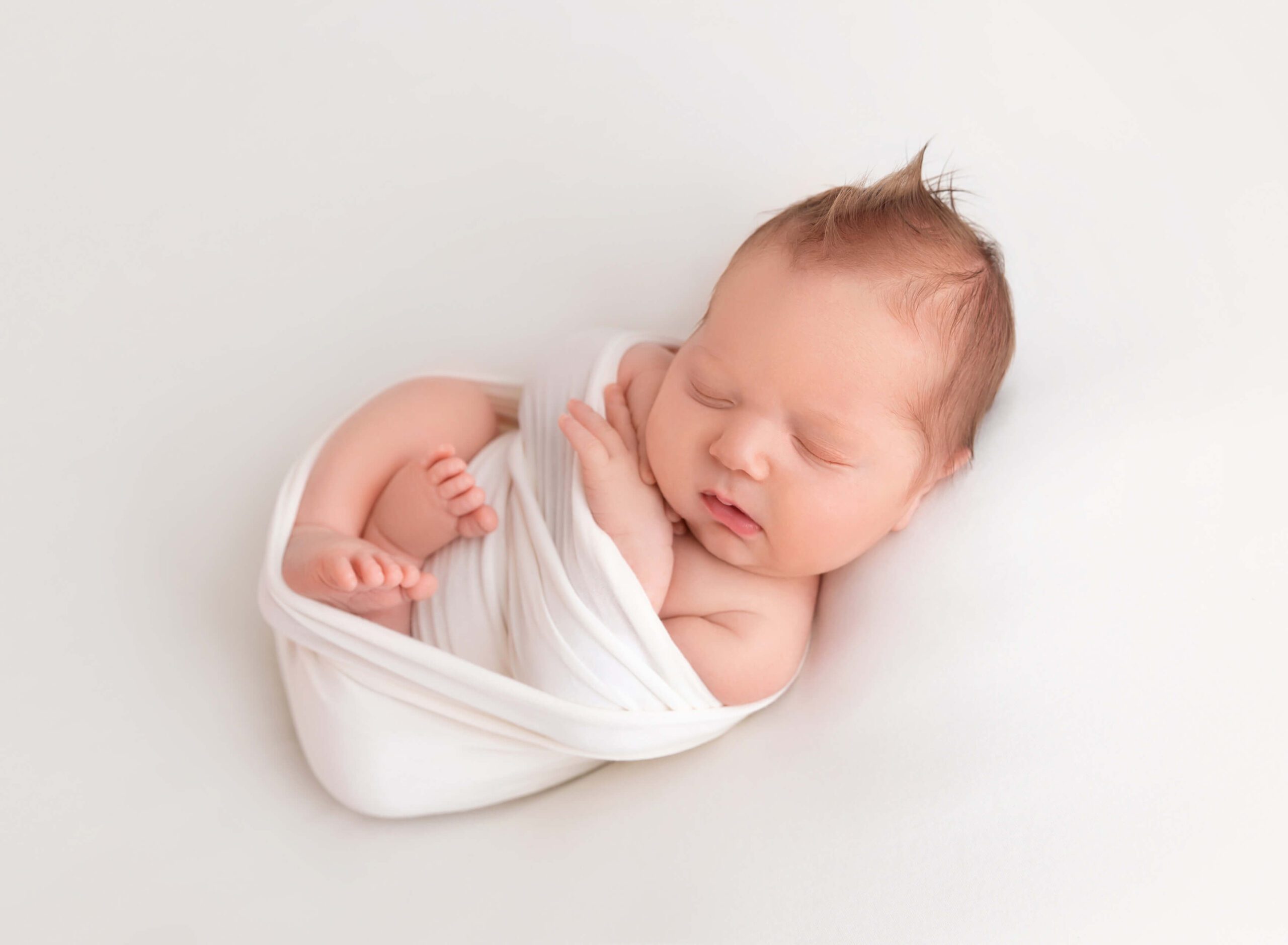 A sleeping newborn baby in a white swaddle on a white bed