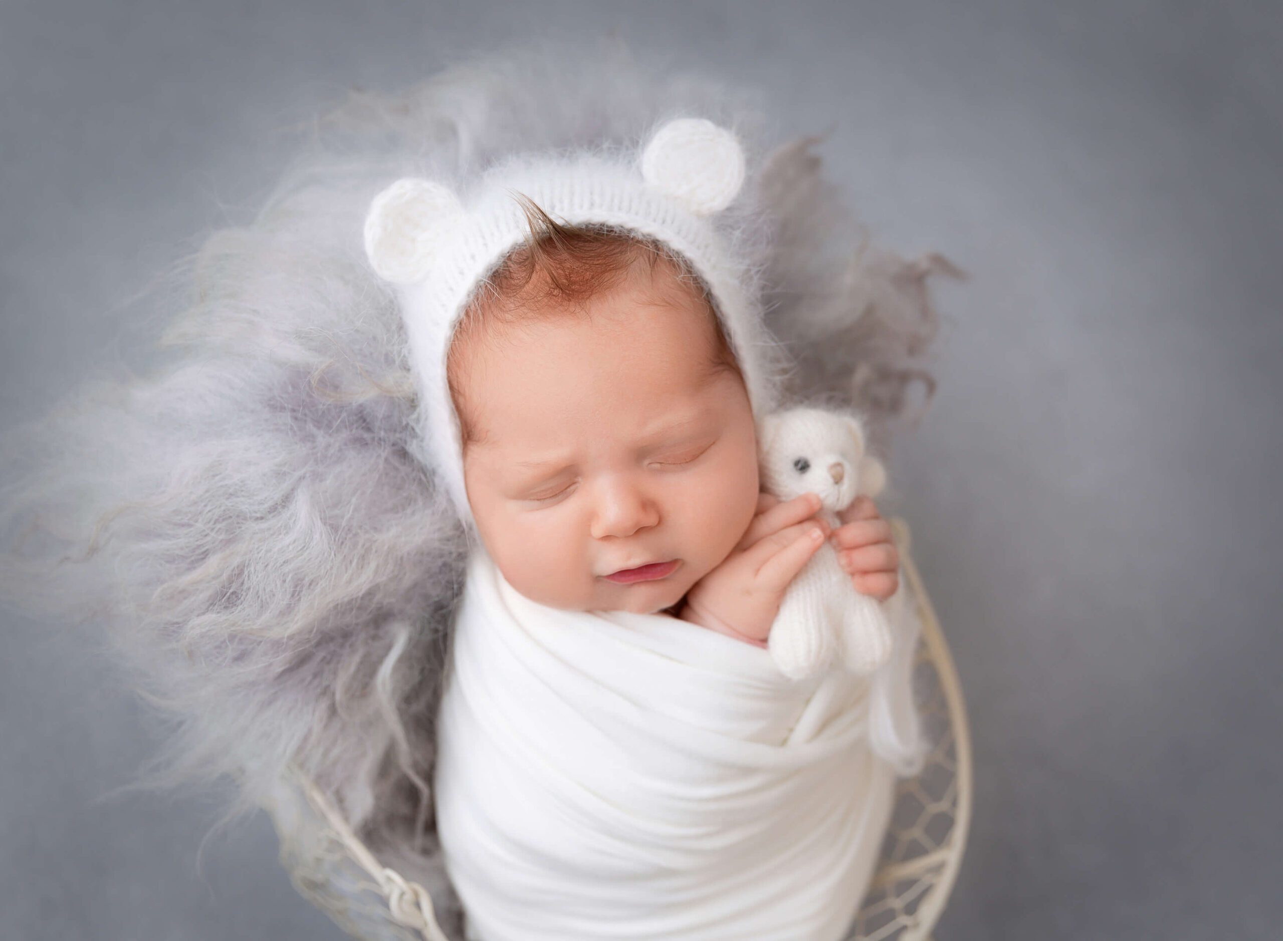 A newborn with red hair sleeps in a white swaddle in a basket holding a white knit teddy after visiting baby boutiques in new york