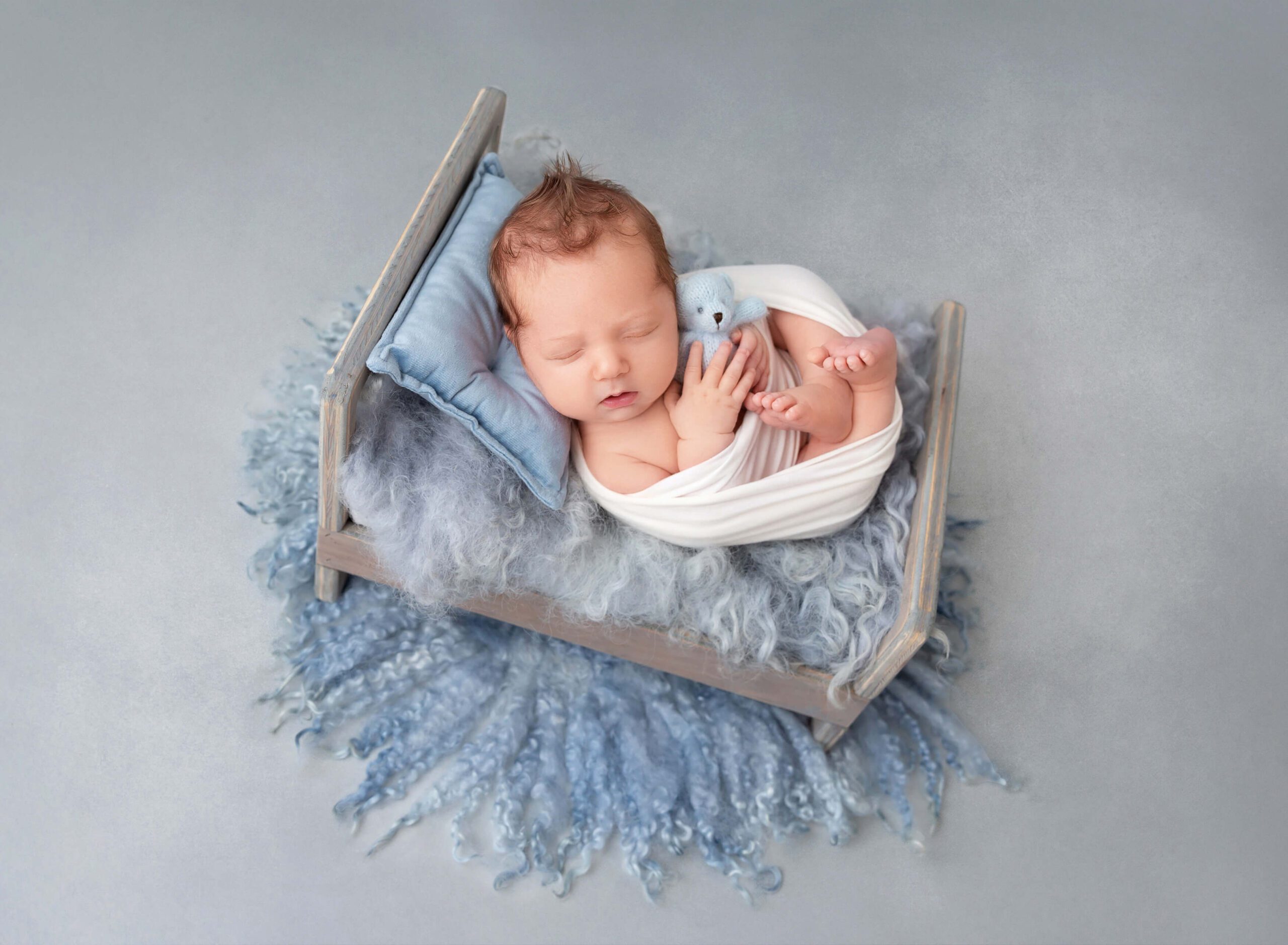 A newborn baby with red hair sleeps in a tiny wooden bed holding a blue teddy bear after visiting baby boutiques in new york