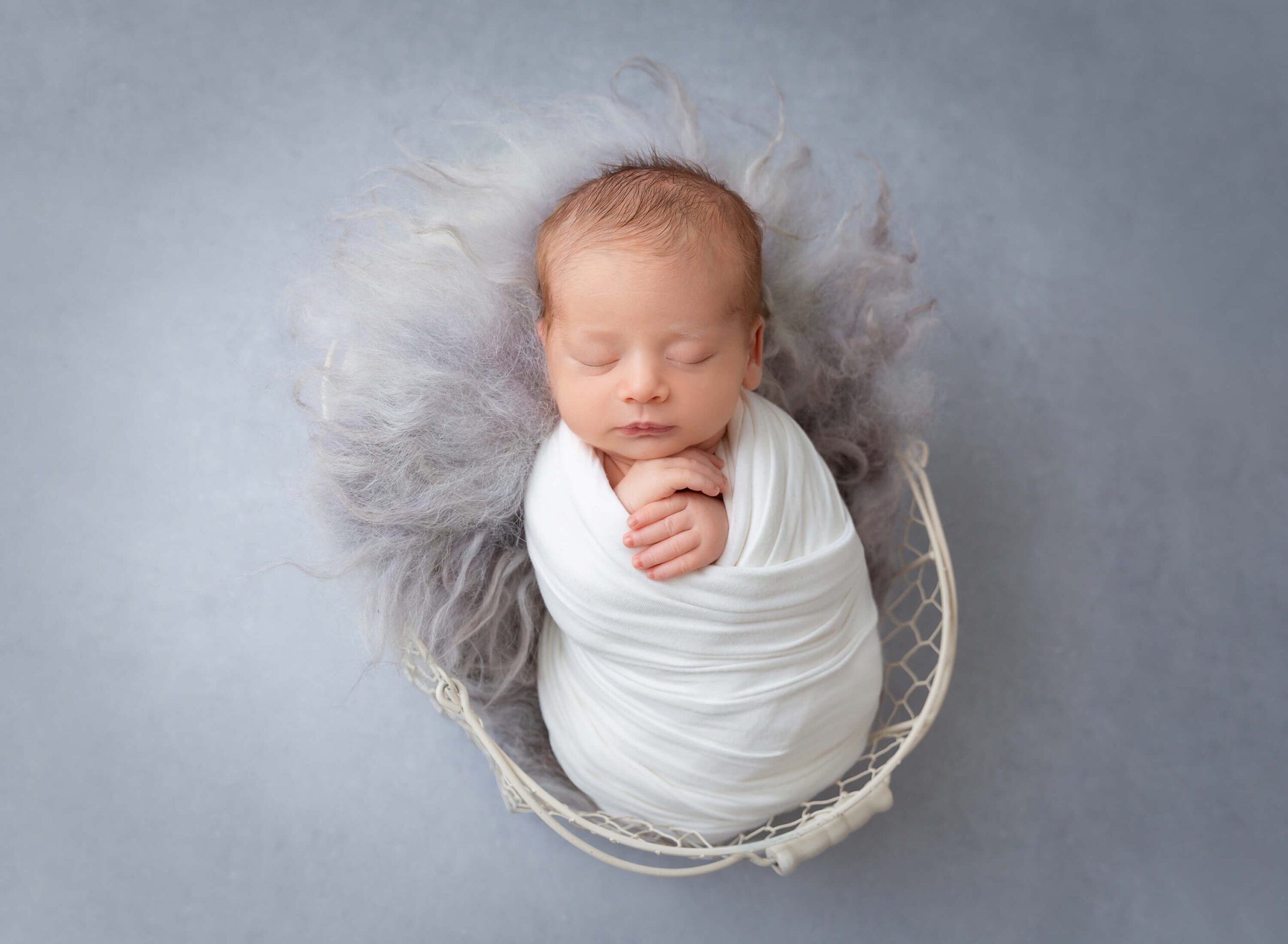 A newborn baby sleeps in a basket in a white swaddle in a studio