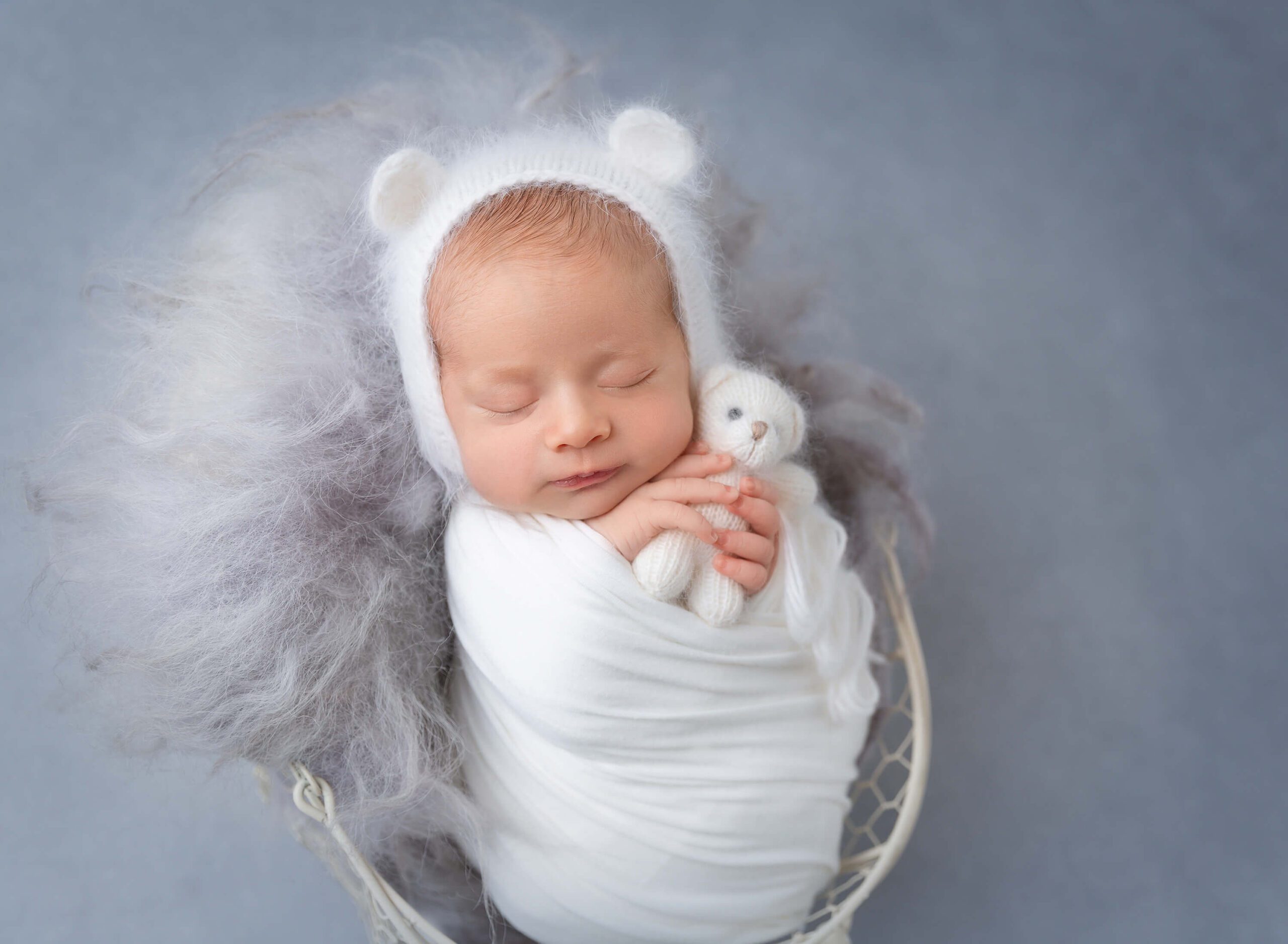 A newborn holds a white knit bear while sleeping in a basket in a knit bear bonnet