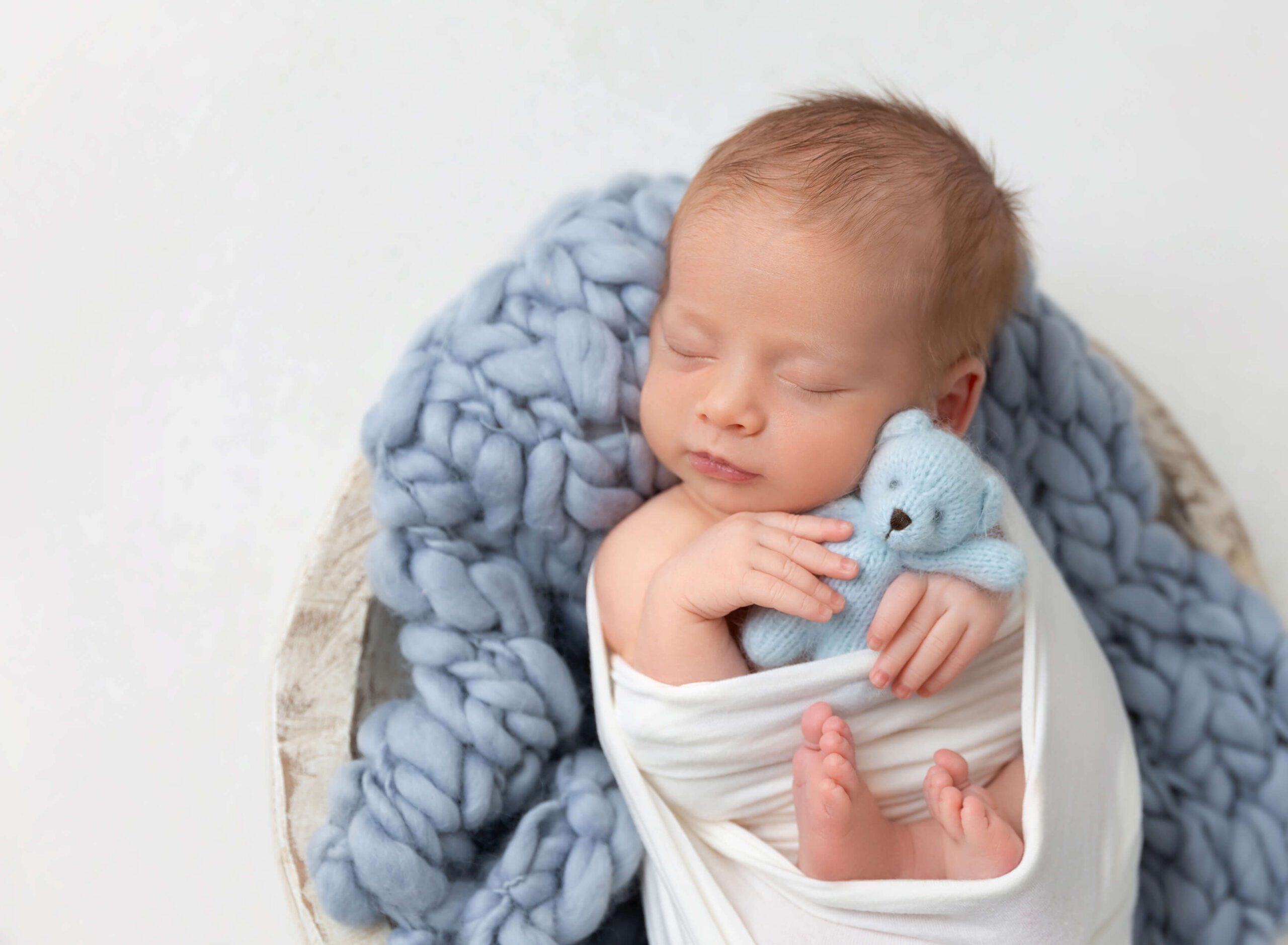 A newborn baby cuddles a blue knit bear in a wooden bowl in a studio while sleeping after meeting a lactation consultant in New York