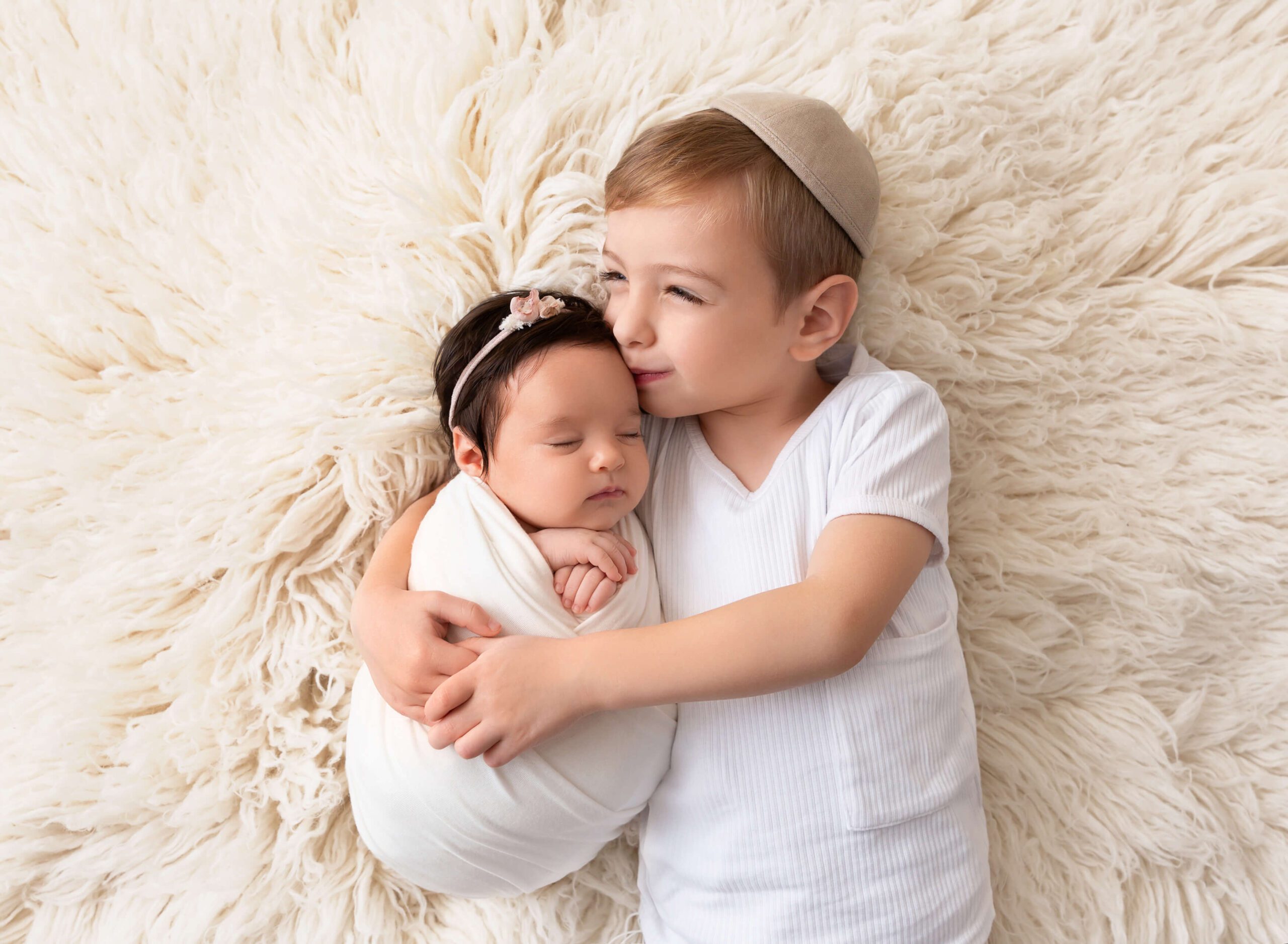 A smiling toddler boy in a white shirt kissing his sleeping newborn sister on a fur blanket after some parenting classes in new york