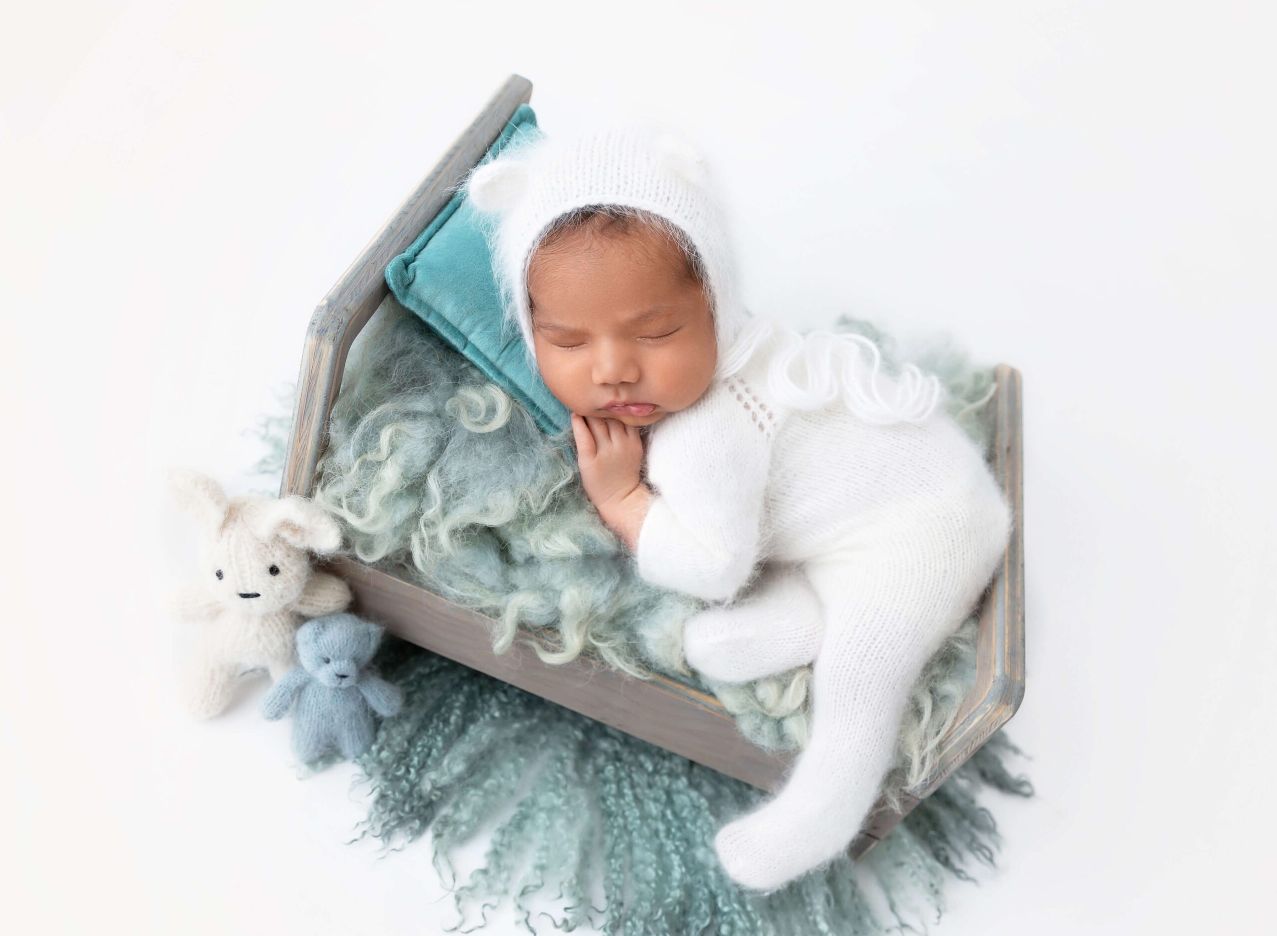 A baby sleeps on a tiny wooden crib in a studio in a white knit onesie and matching bear ear bonnet after visiting pediatric dentists in new york