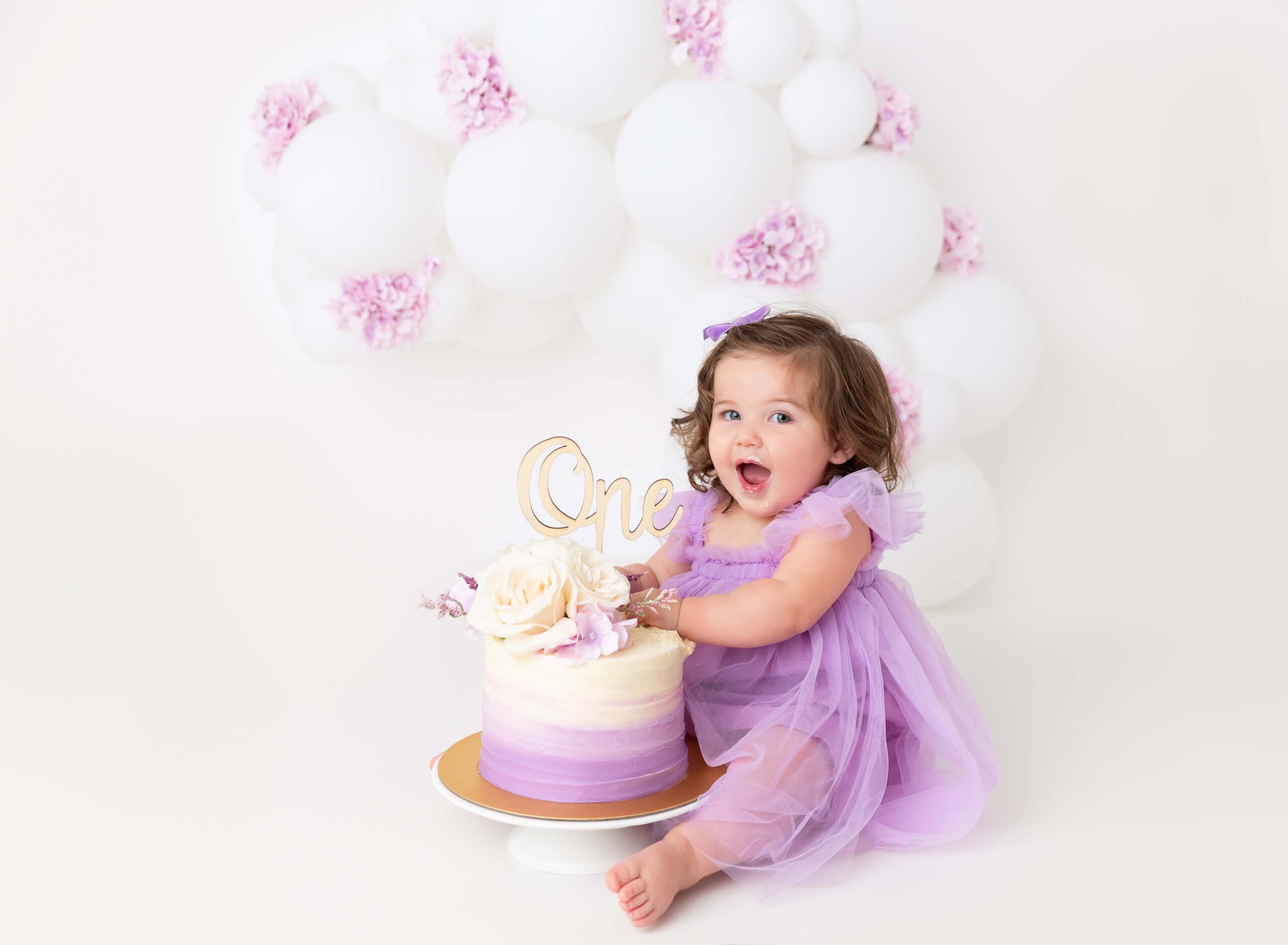 A happy toddler in a purple dress shoves her hands in a matching cake in a studio