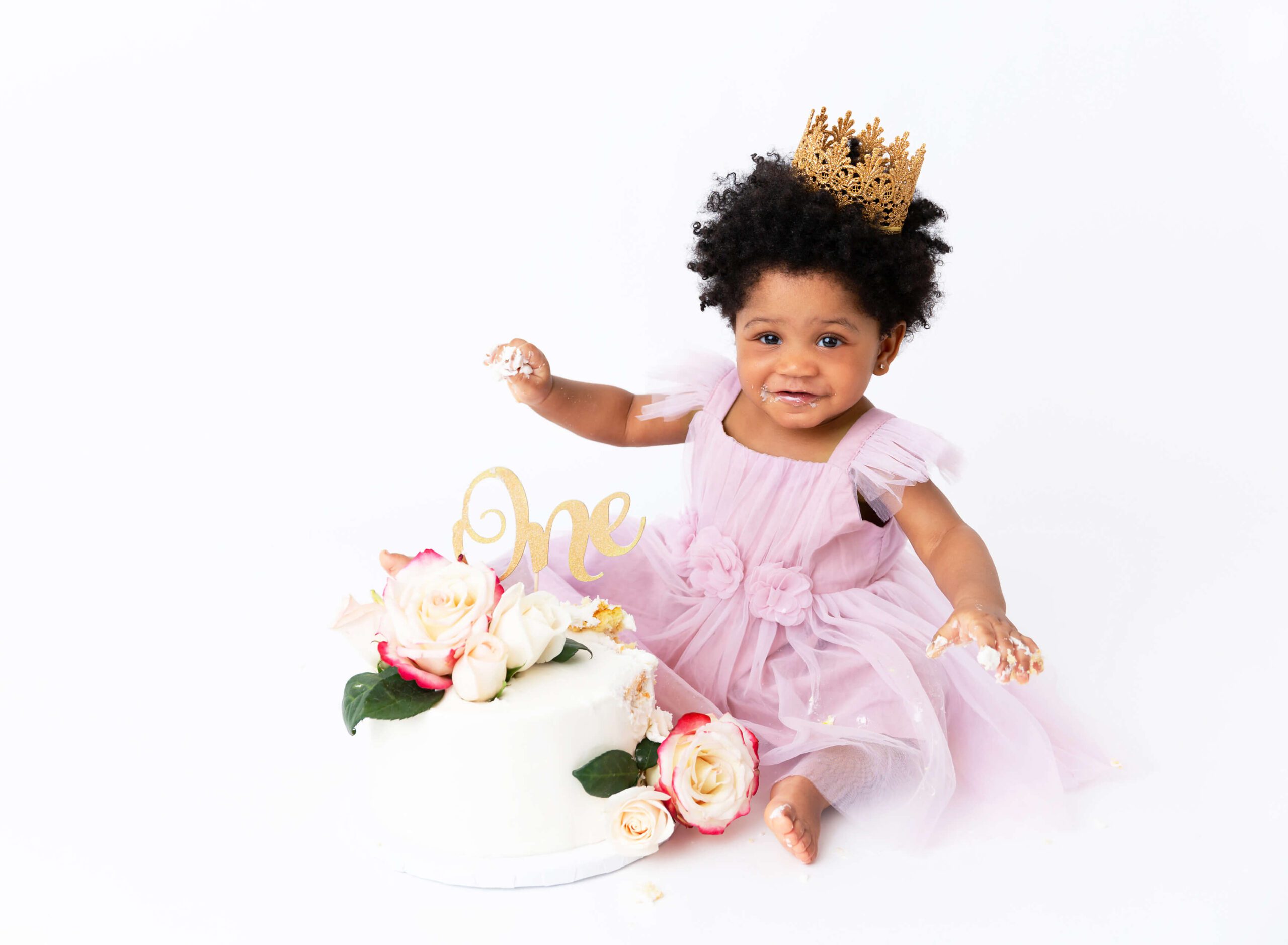 A baby in a pink dress plays with a white birthday cake with flowers in a studio after meeting pediatricians in new york