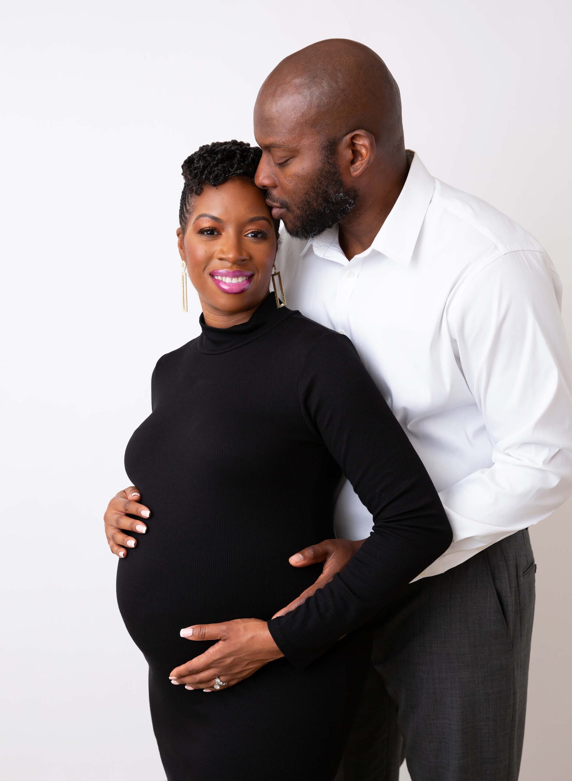 A man leans over kissing his pregnant wife in a black maternity one piece maternity suit after prenatal yoga in new york