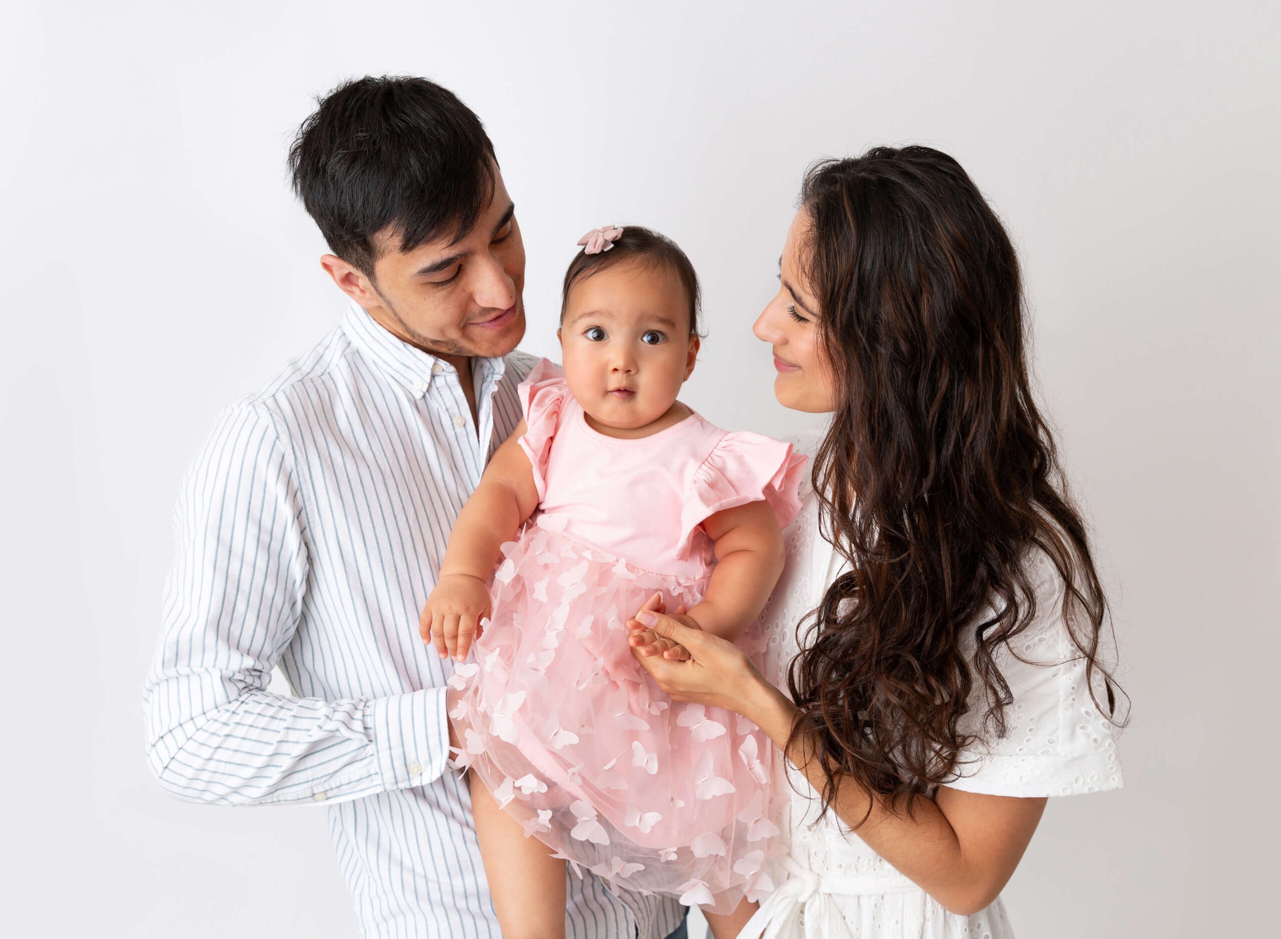 A happy mom and dad stand in a studio holding their baby girl in a pink dress