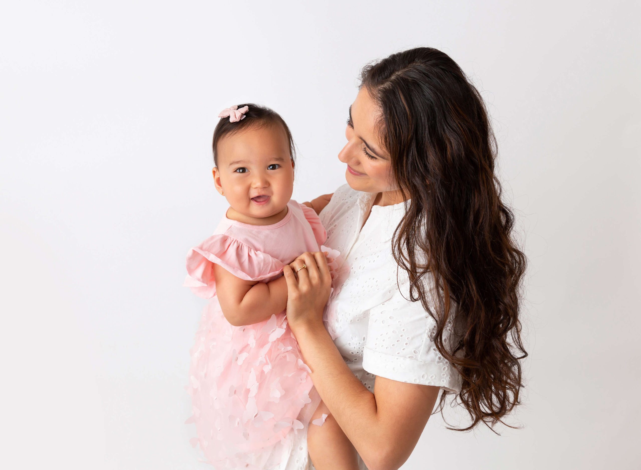 A happy baby in a pink dress sits in mom's arms in a studio after some Mommy and Me Classes in New York