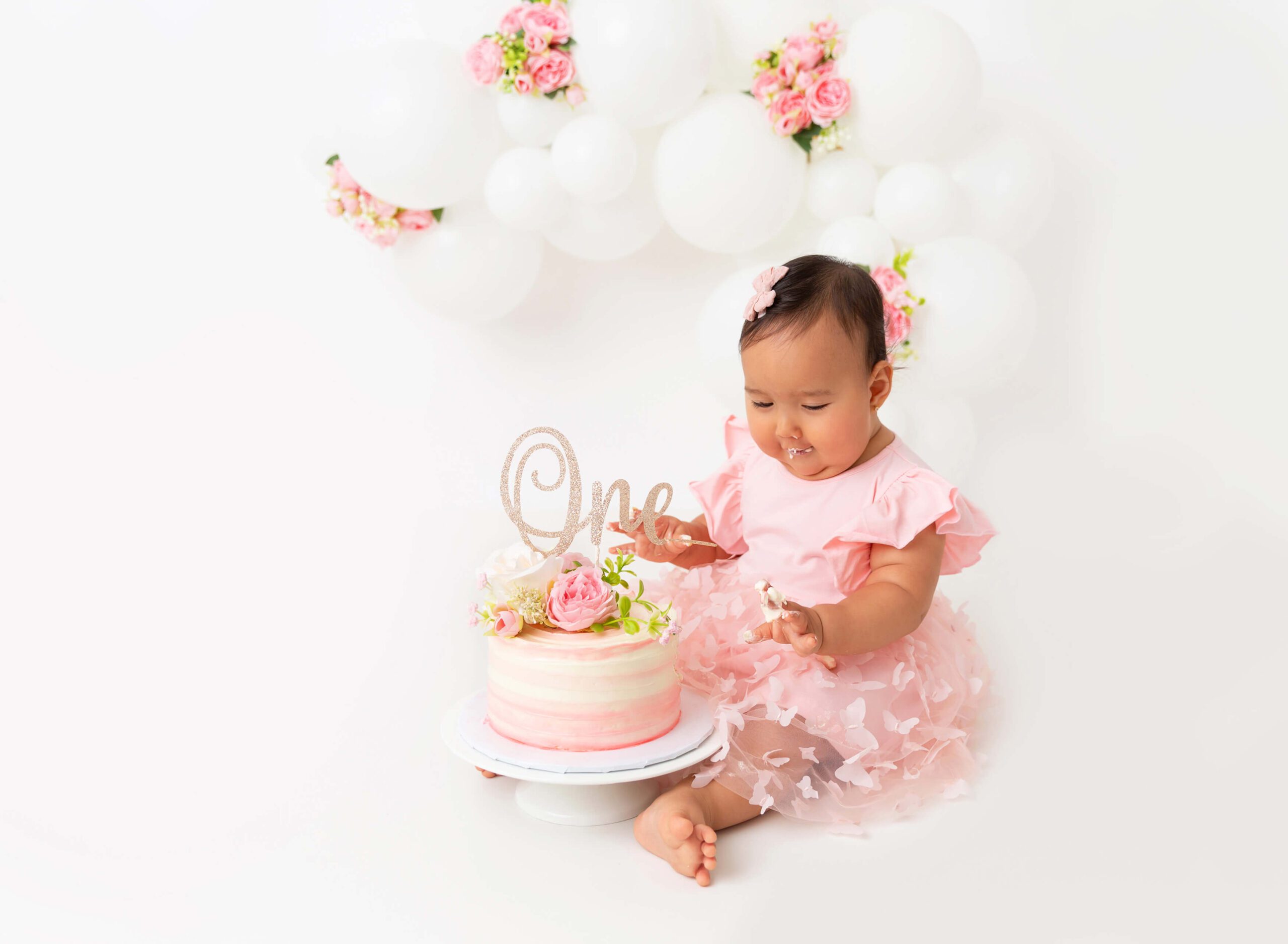 A happy baby girl in a pink dress eats cake of her fingers in a studio after some Mommy and Me Classes in New York