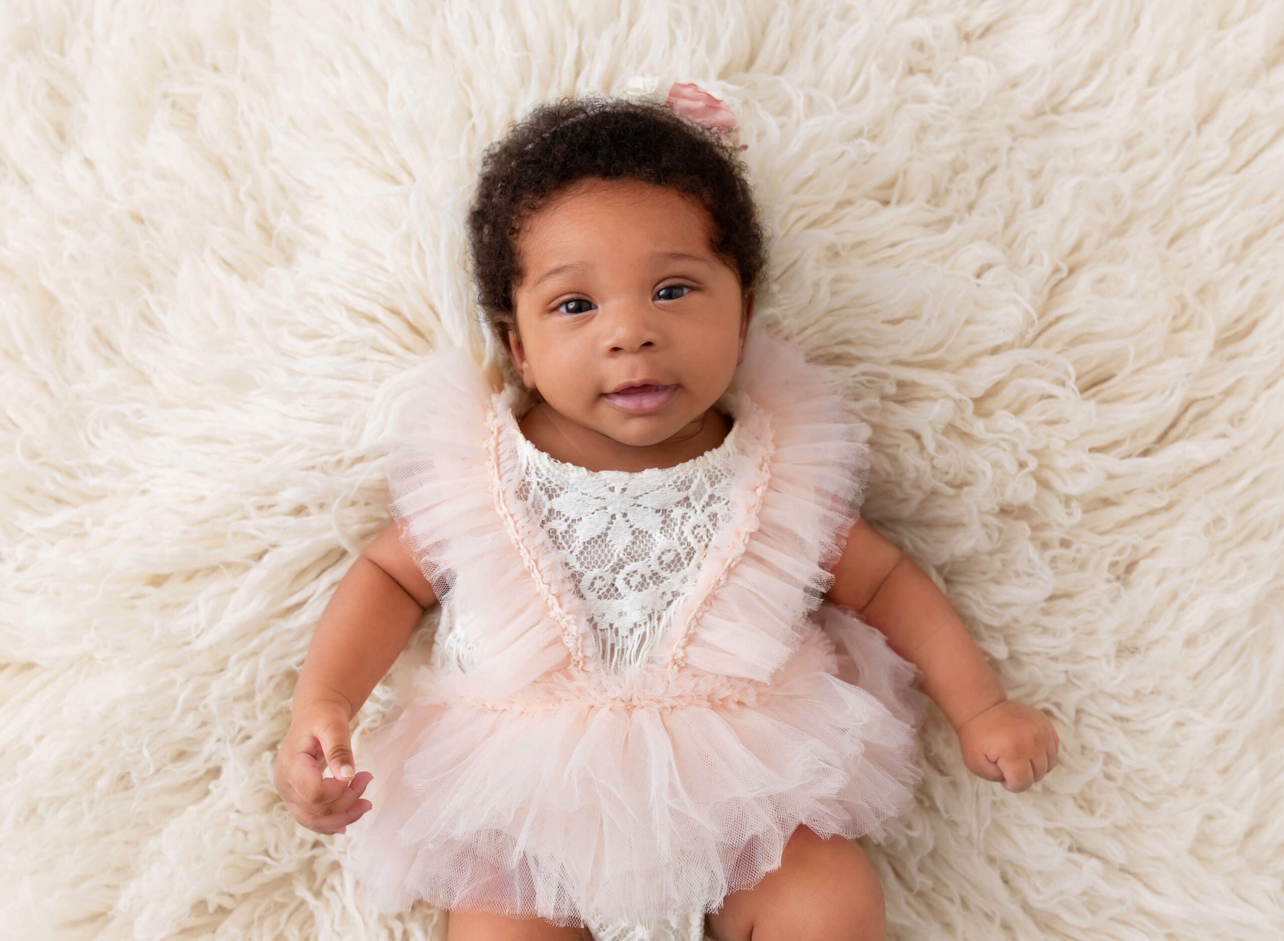 A happy baby girl in a pink dress lays on a white shag balnket