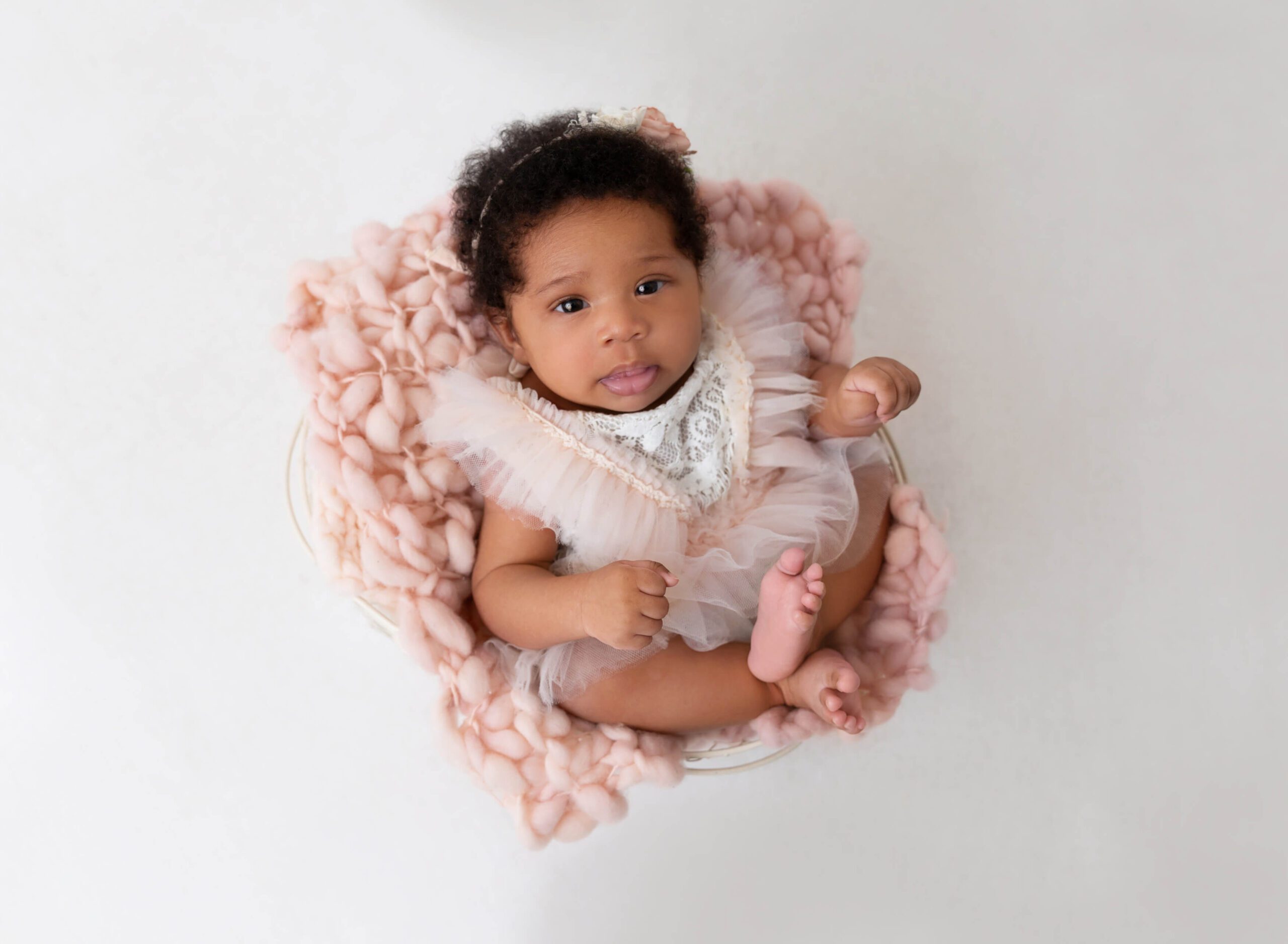 A happy newborn baby in a tule and lace dress sits awake in a bucket on pink blanket after visiting Peanut and Honey