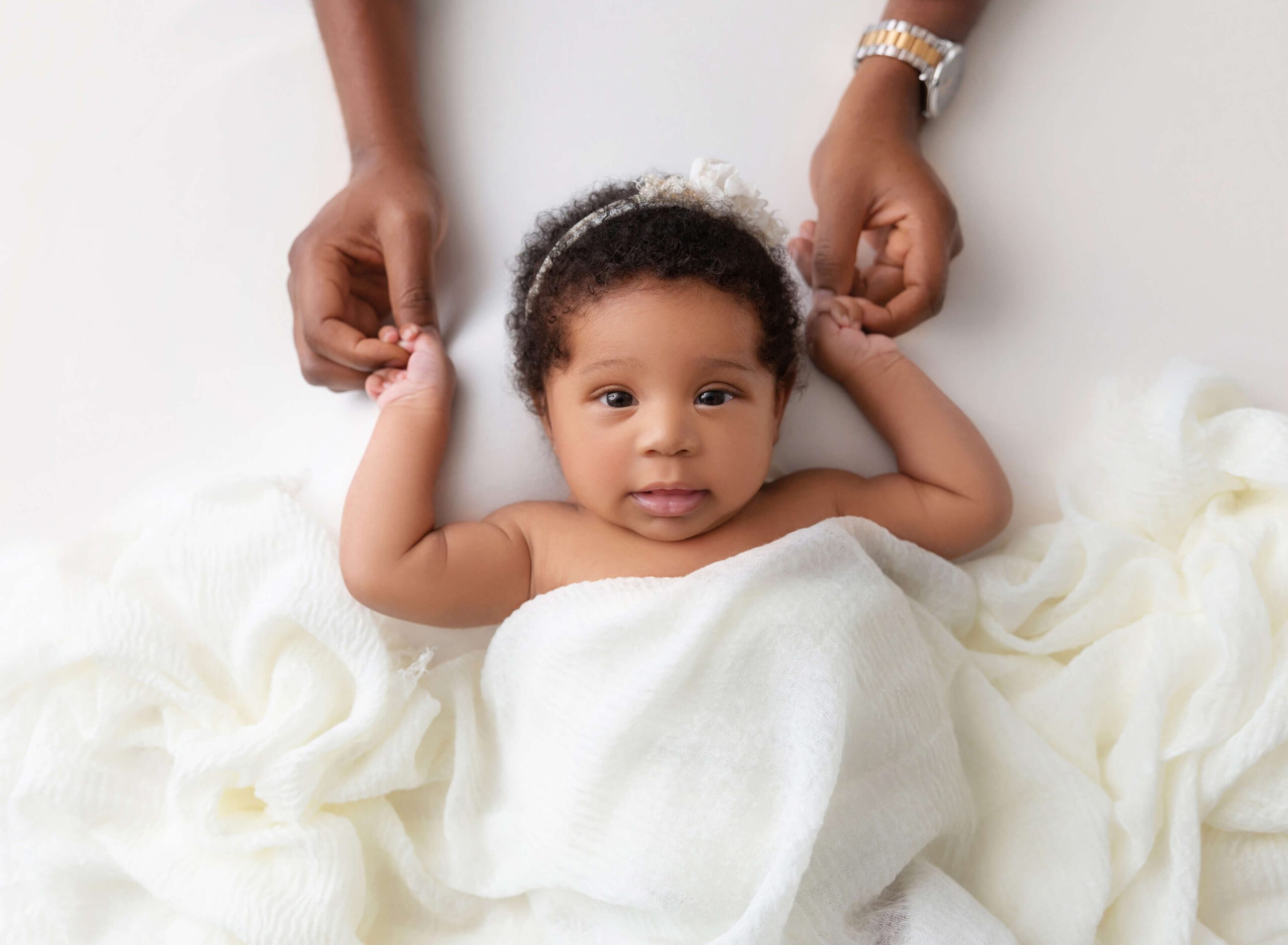 A newborn baby girl lays under a white blanket holding mom's hands above her head with eyes open after visiting Peanut and Honey