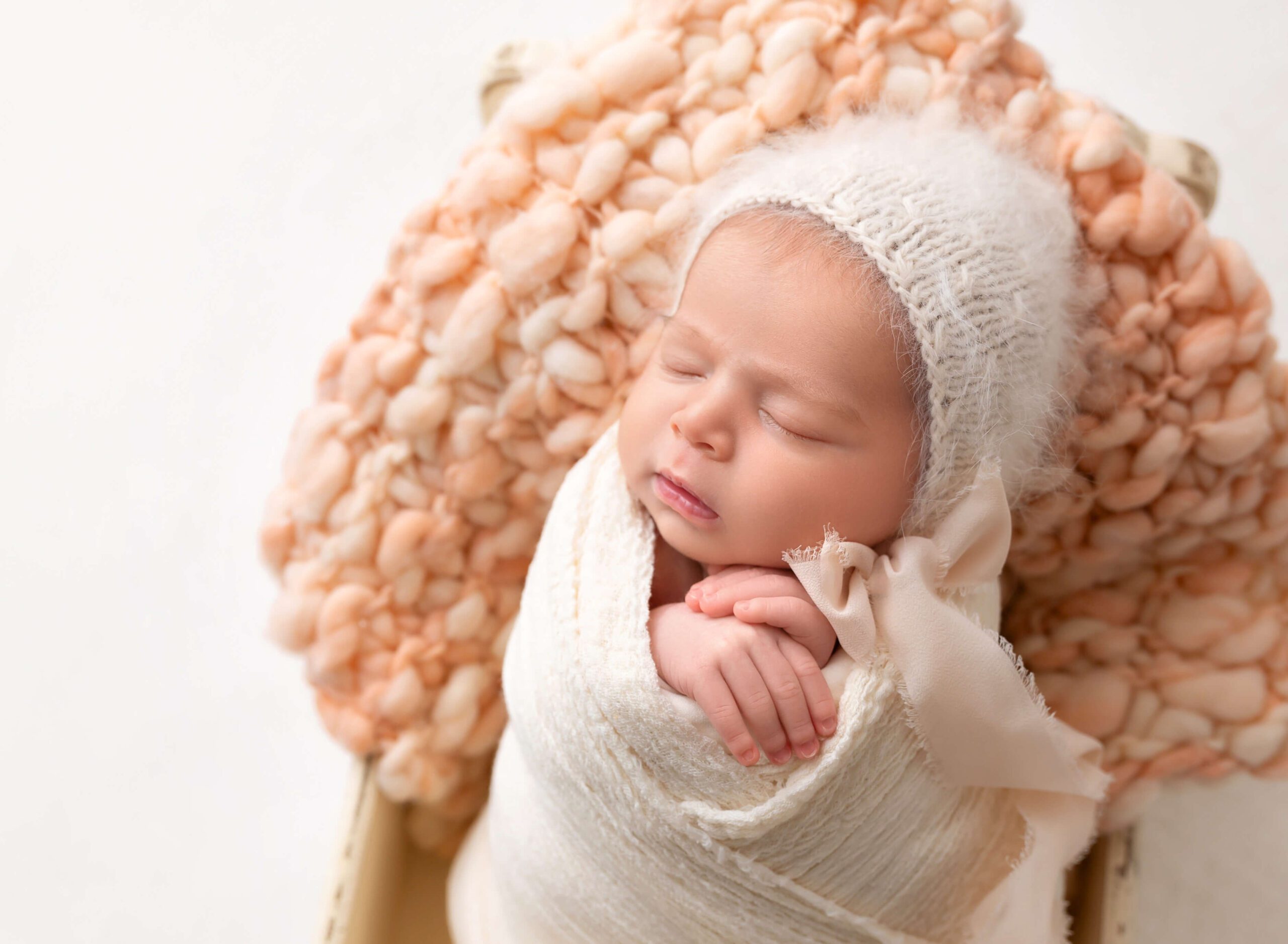 A newborn baby sleeps in a white swaddle and knit beanie in a wooden crib on a pink blanket after meeting babysitters new york