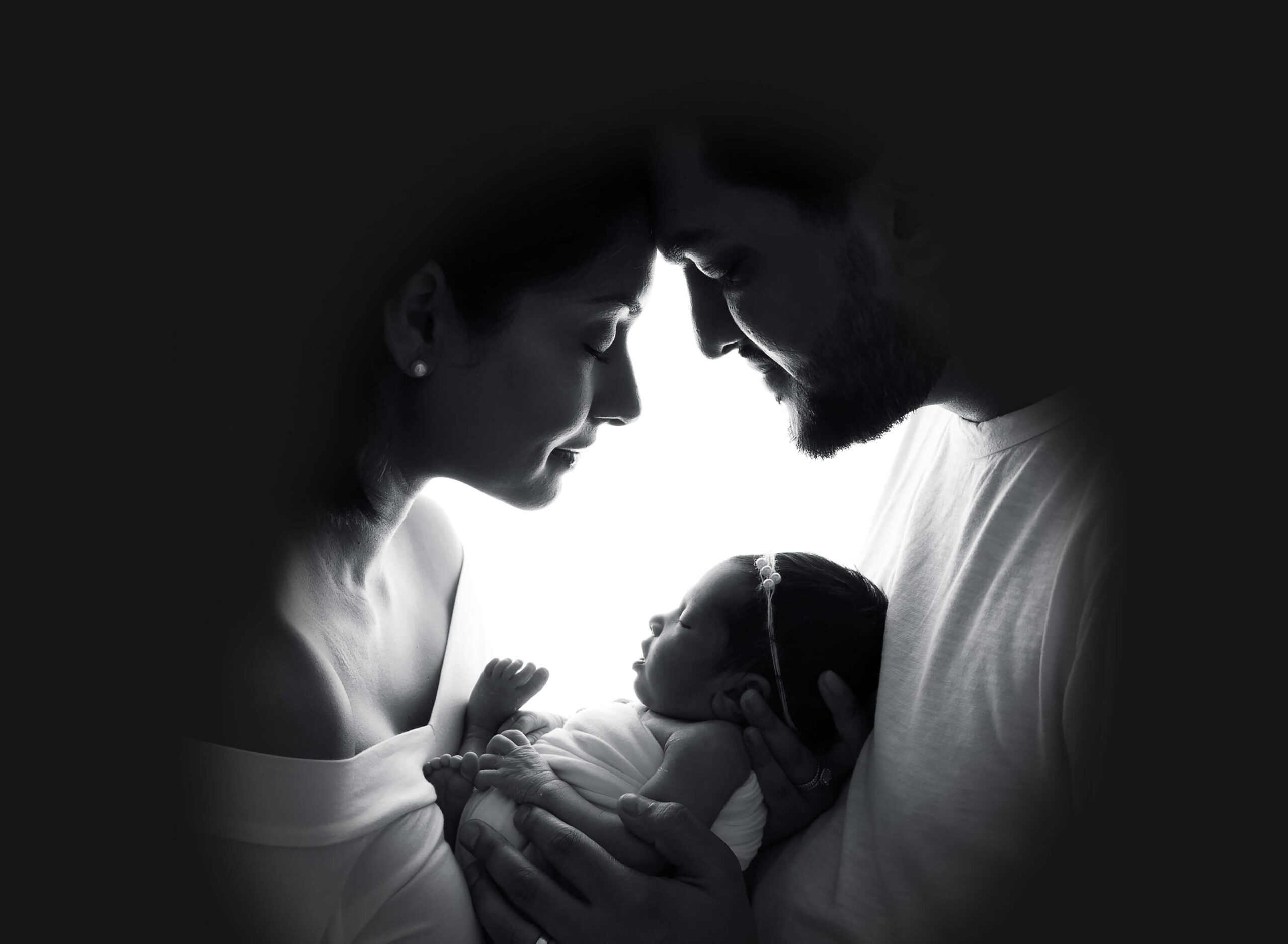 A newborn baby girl sleeps in mom and dad's hands between them against their chests as they touch foreheads