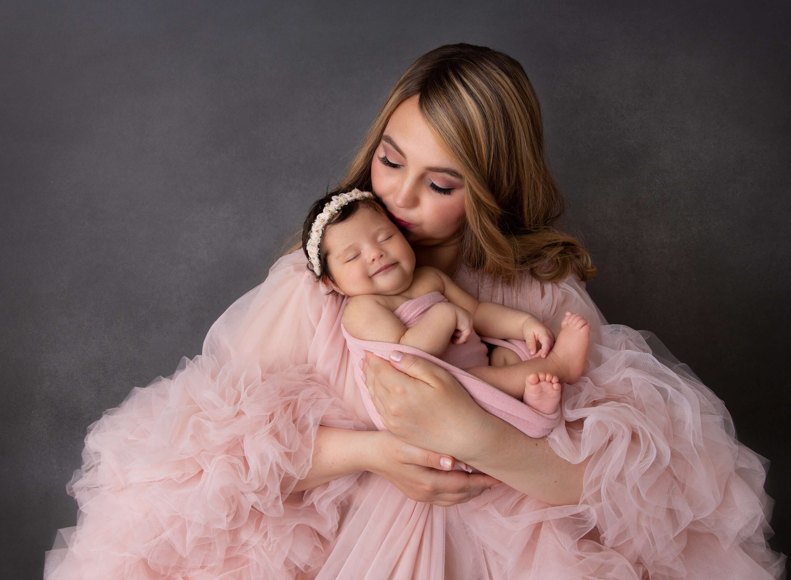 A smiling newborn baby girl sleeping in mom's hands while being kissed in a studio after visiting maternity stores in new york