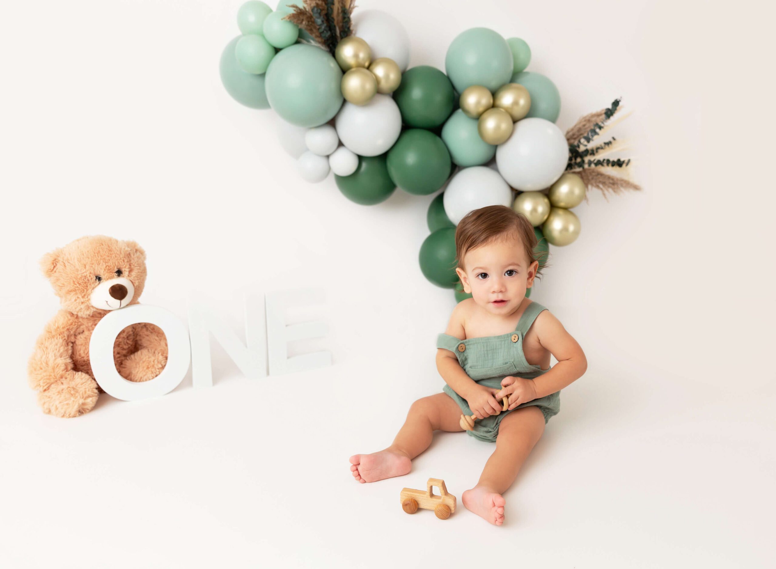 A happy baby boy in green overalls plays with some wooden toys in a studio