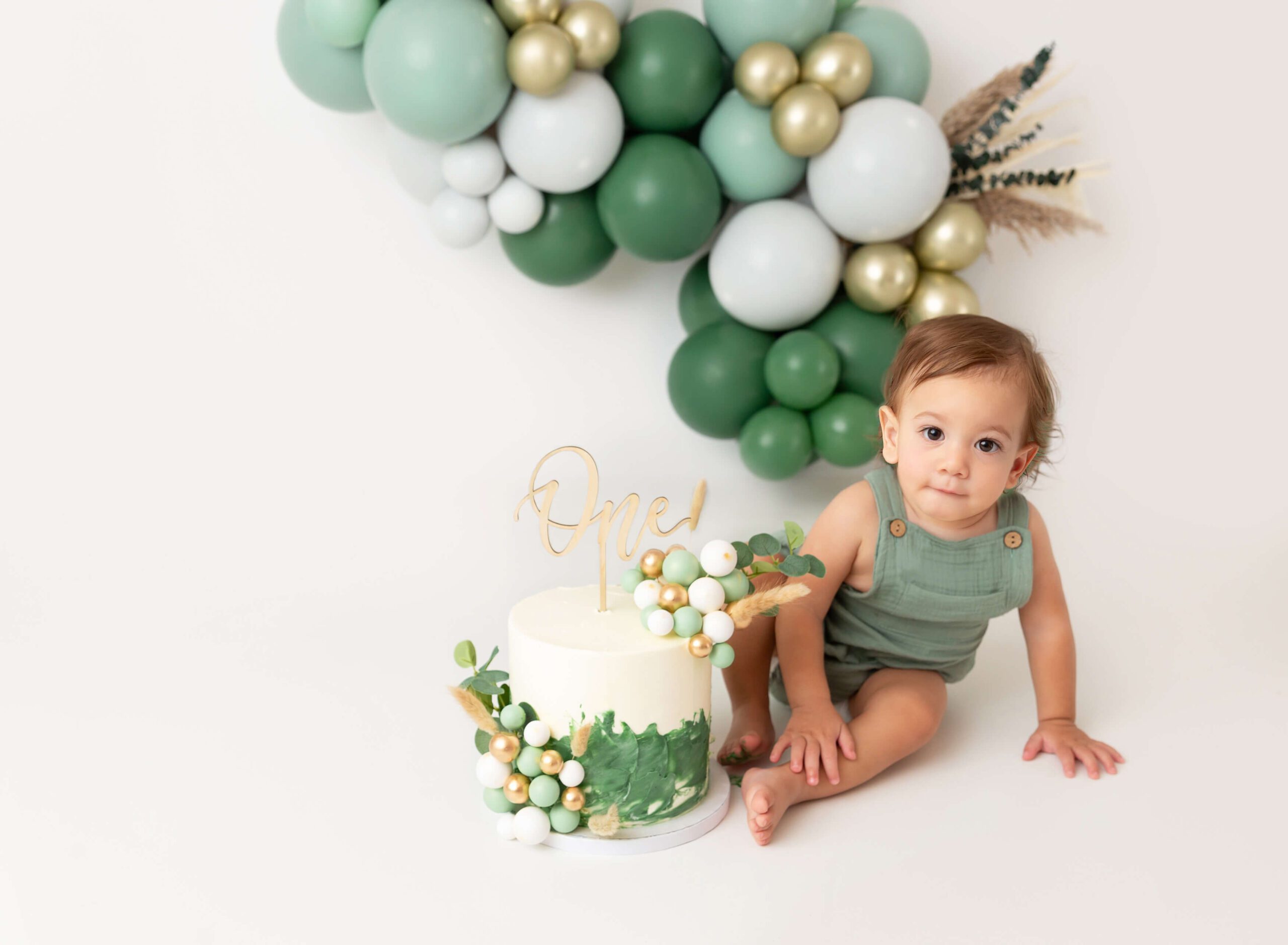 A baby in green overalls sits behind a birthday cake with balloons after some music classes for babies in nyc