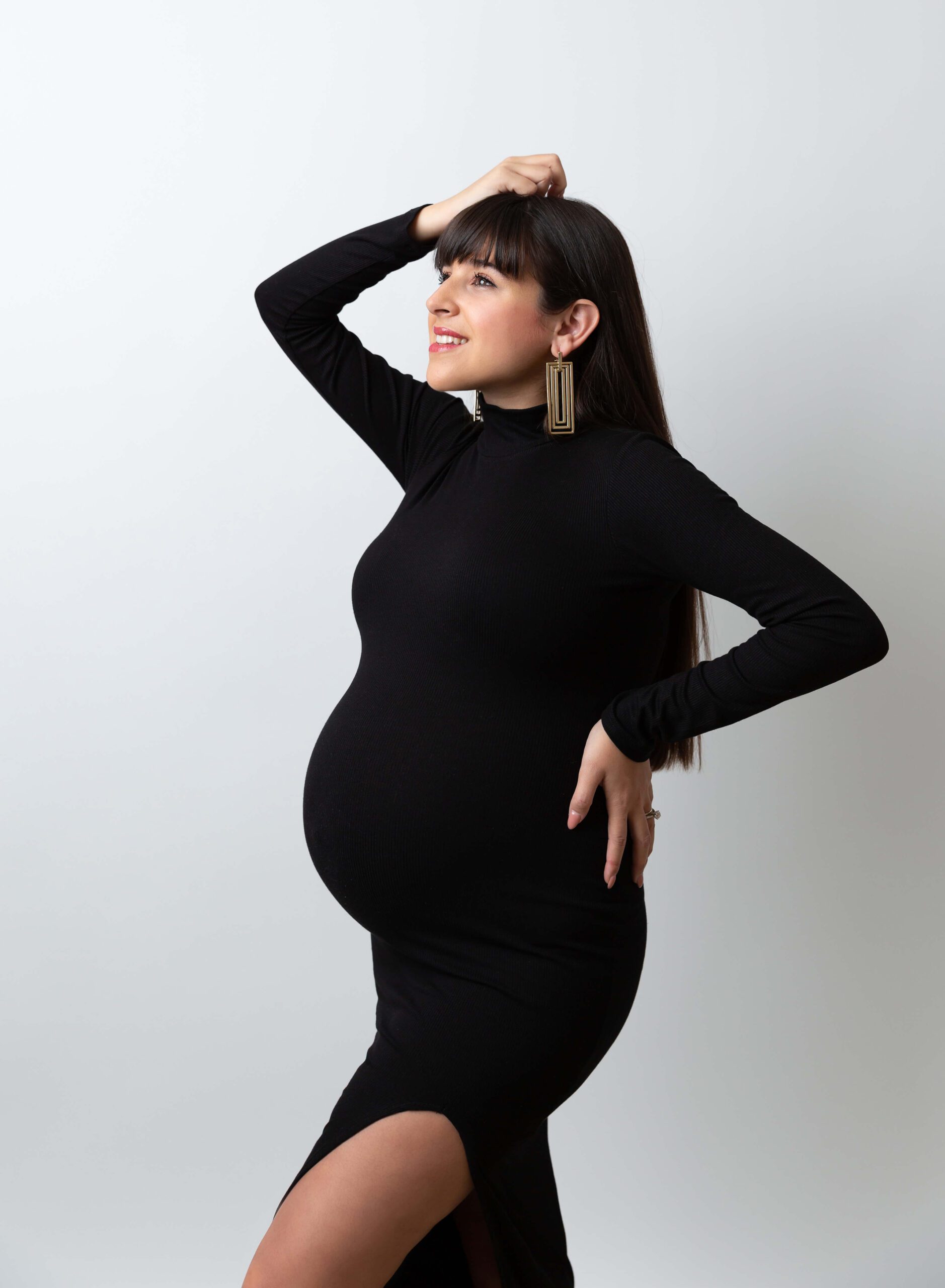 A happy mom to be in a tight black maternity dress stands in a studio with a hand on her hip and in her hair