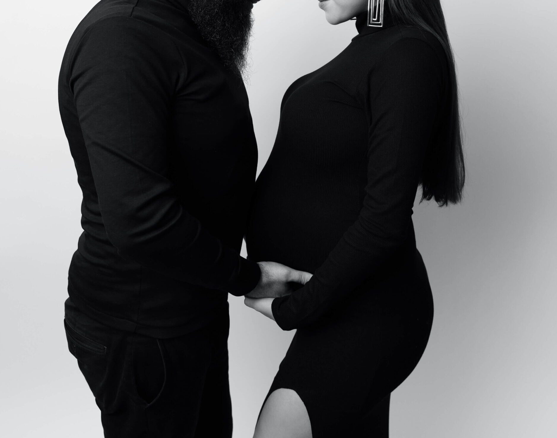 A happy pregnant couple holds hands under the bump in black and white in a studio before mom needs pelvic floor therapy in new york