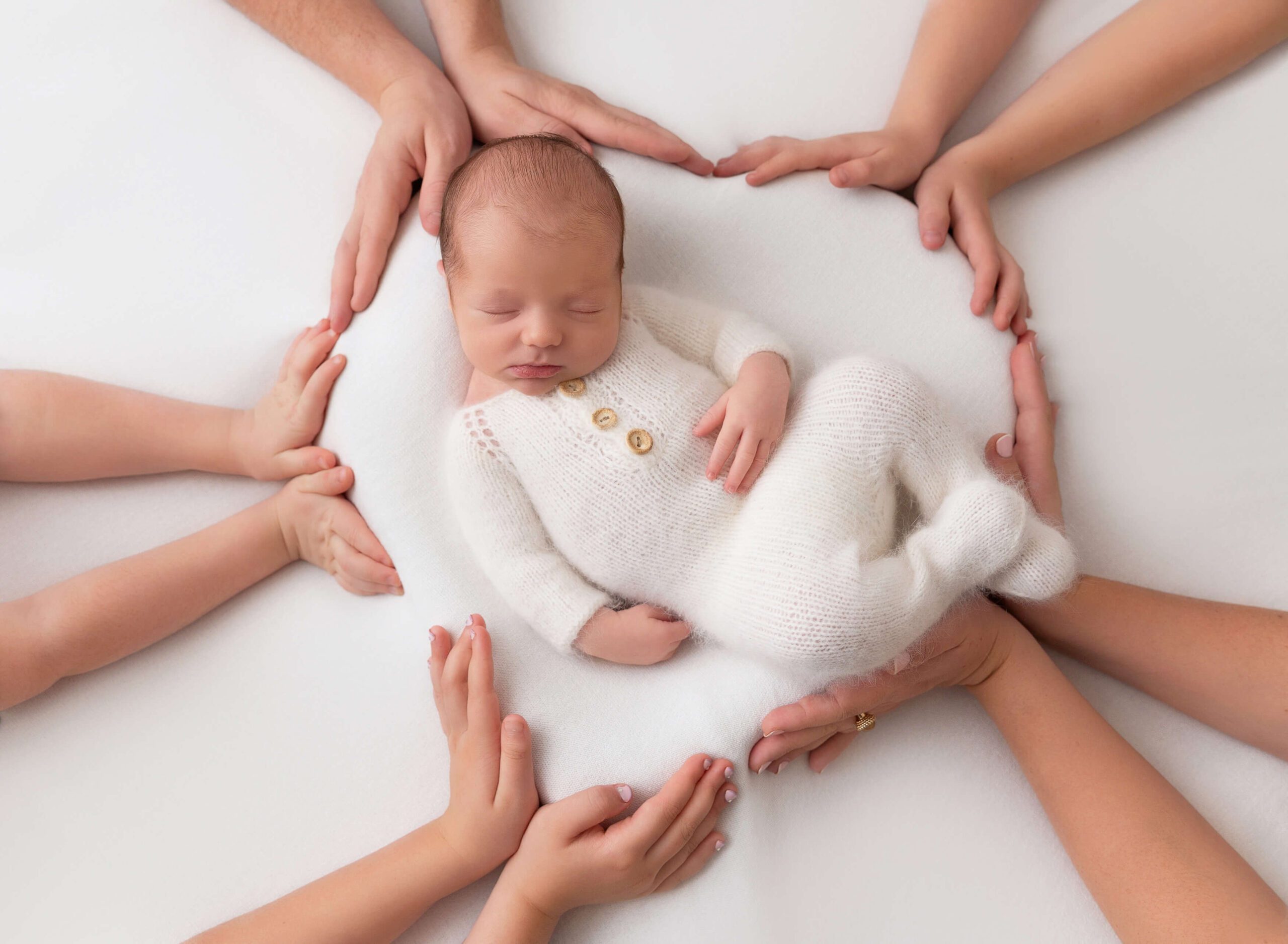 A family holds their hands in a circle around a sleeping newborn baby in a white knit onesie