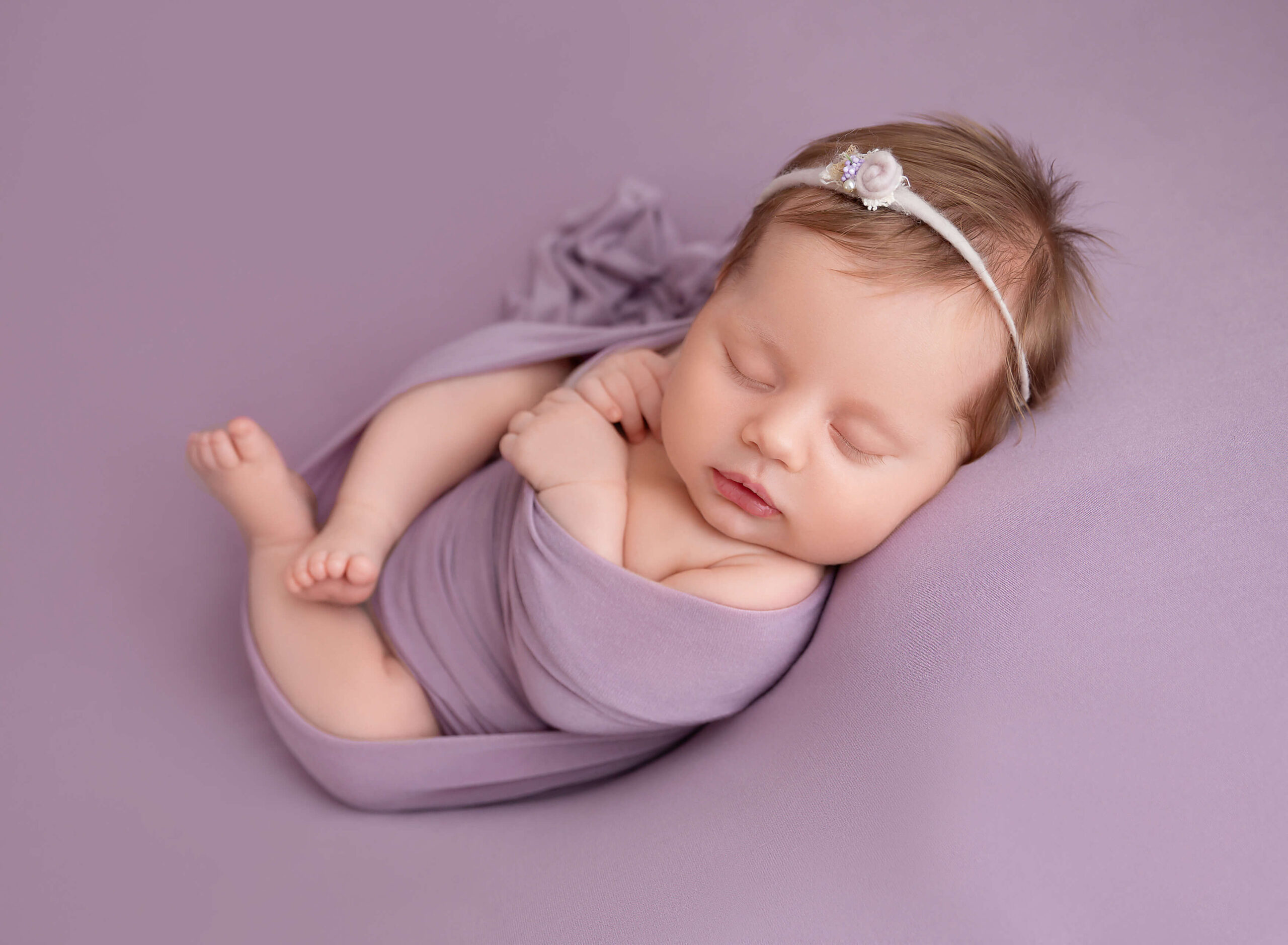 A sleeping newborn baby lays in a purple wrap with a tiny headband