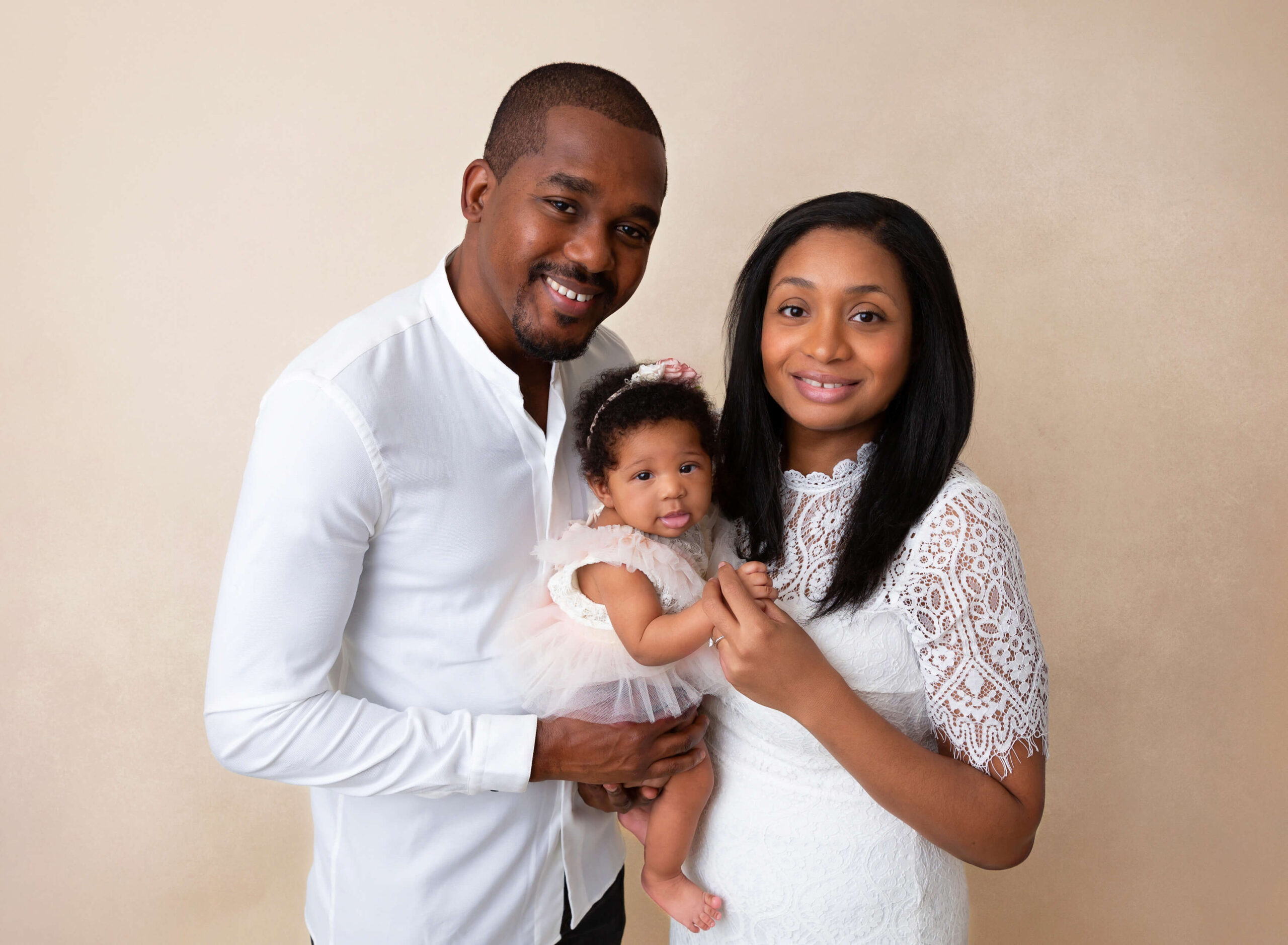 Happy mom and dad stand in a studio holding their baby girl in a white dress