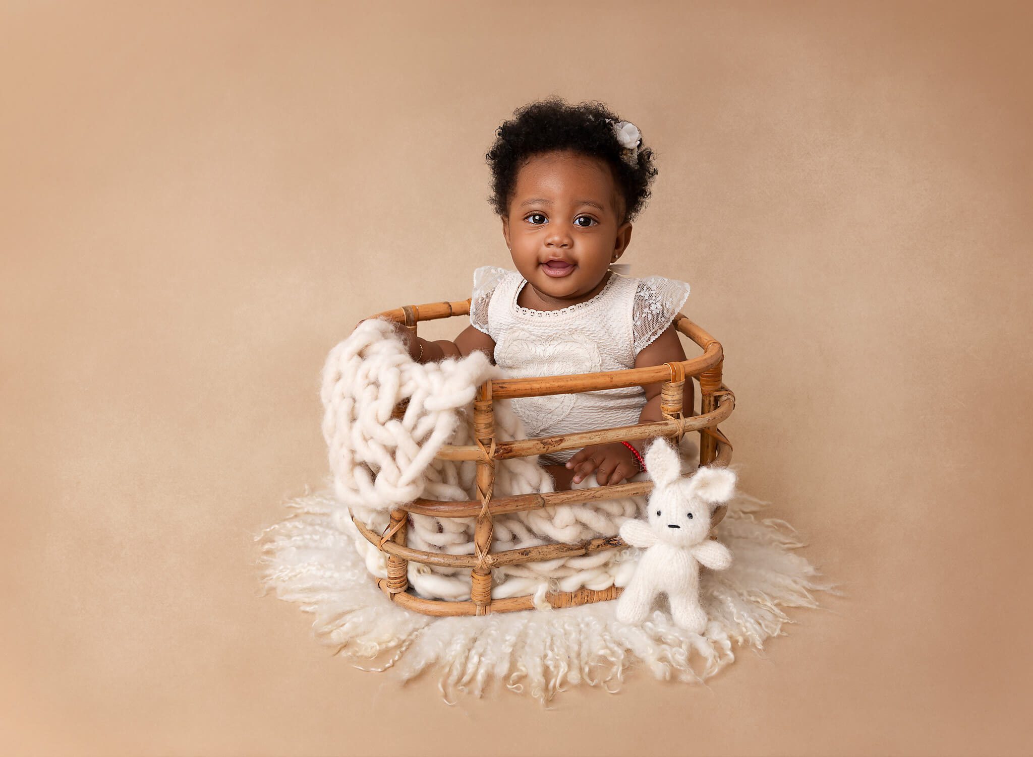 A happy infant girl in a white dress sits in a wicker basket with a white knit bunny after some Baby Classes In NYC