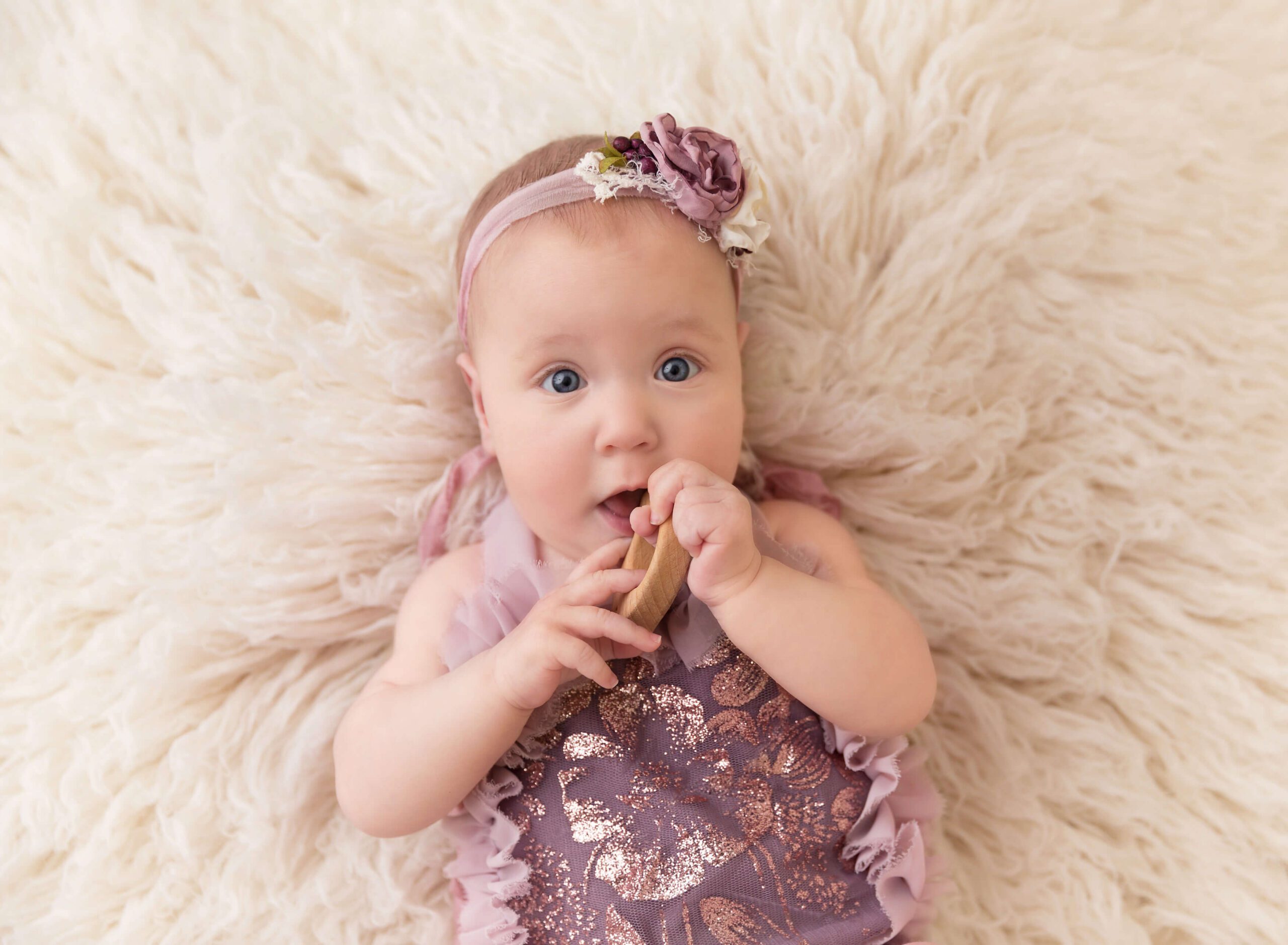 A baby girl lays on her back in a purple dress and matching floral headband with a wooden toy