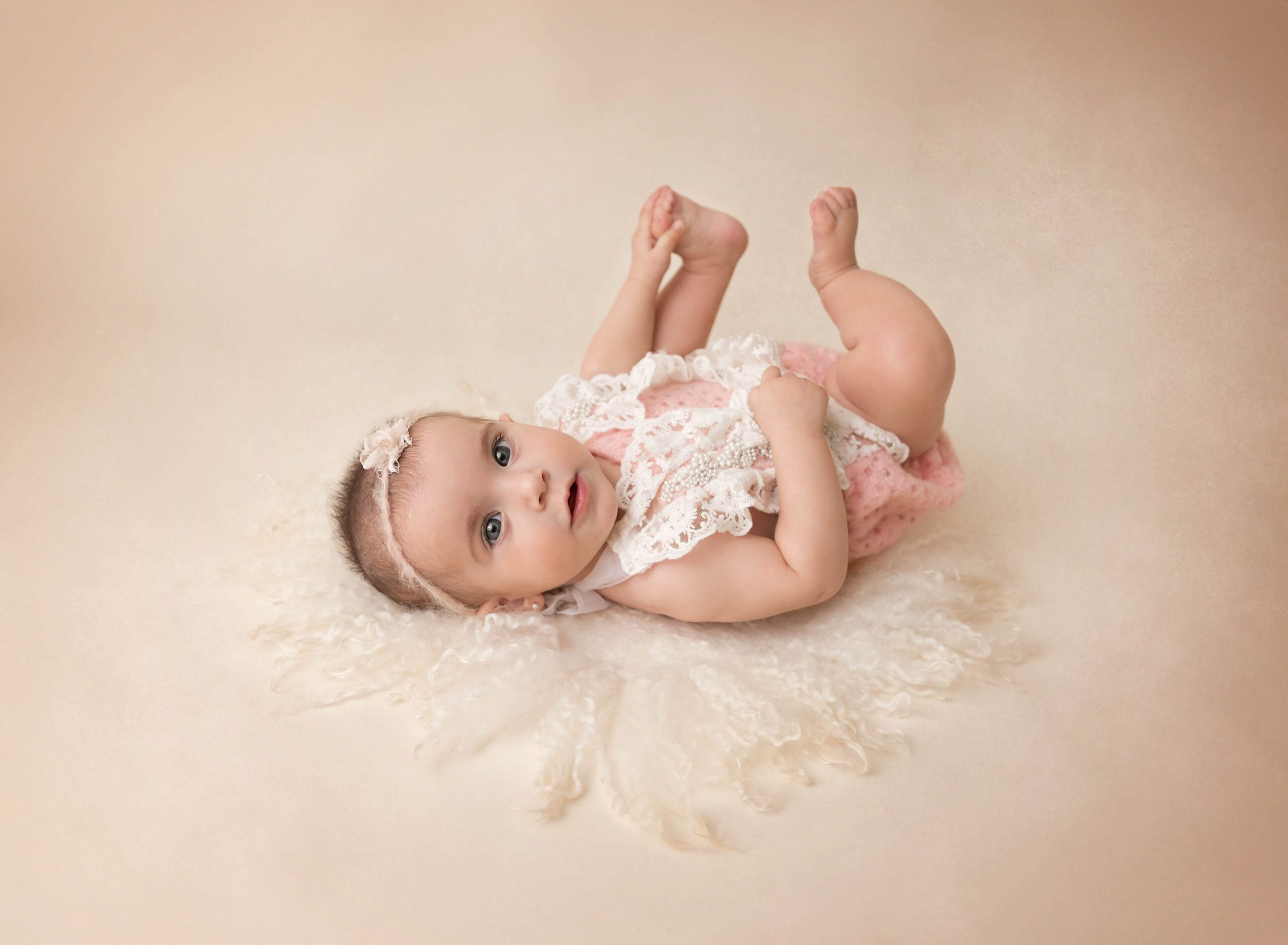 A baby girl in a lace onesie lays on her back touching her feet in a studio after some Baby Yoga In NYC