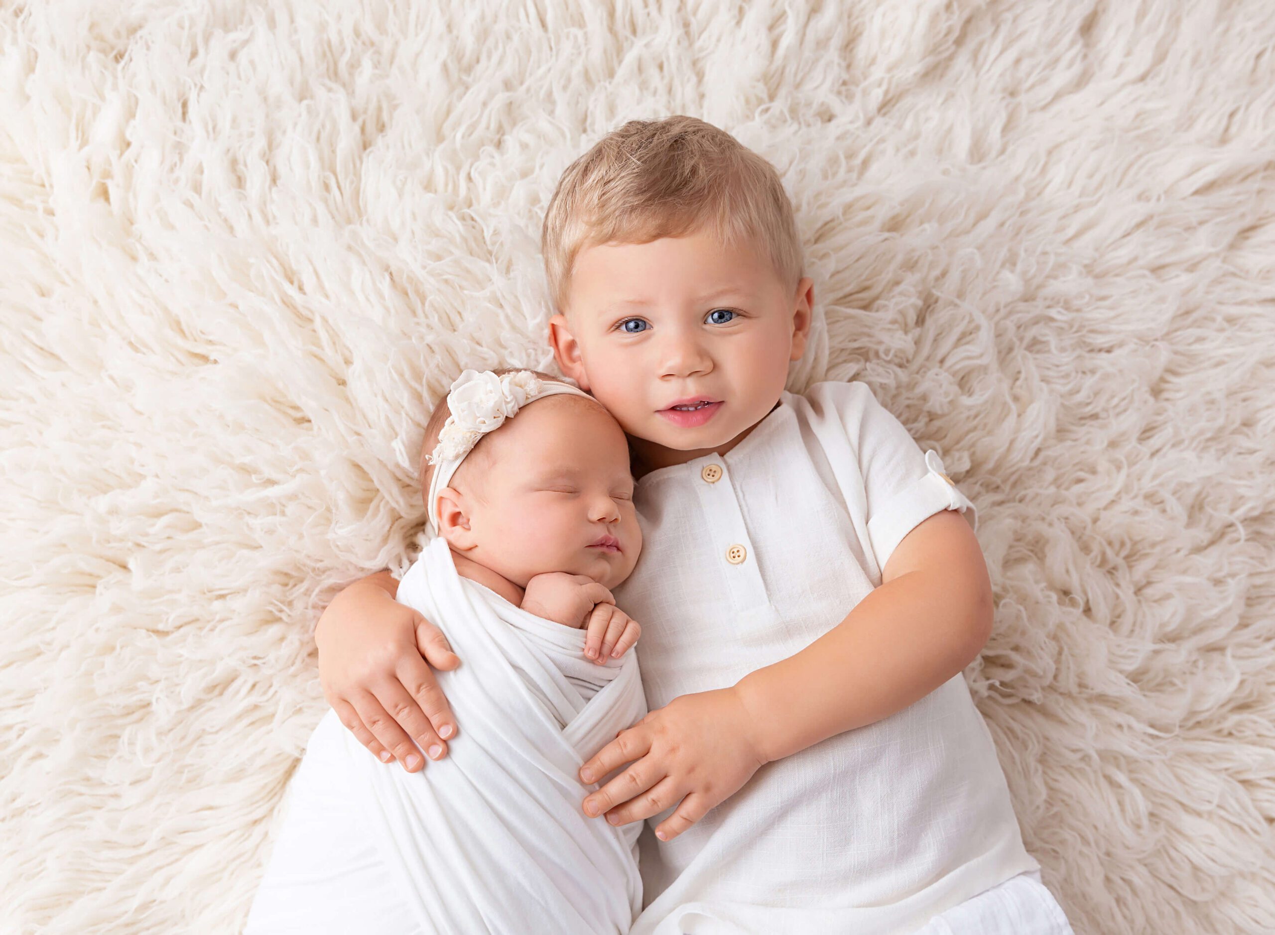 A toddler boy in white snuggles his sleeping newborn sister on a shag blanket after visiting Daycares In NYC