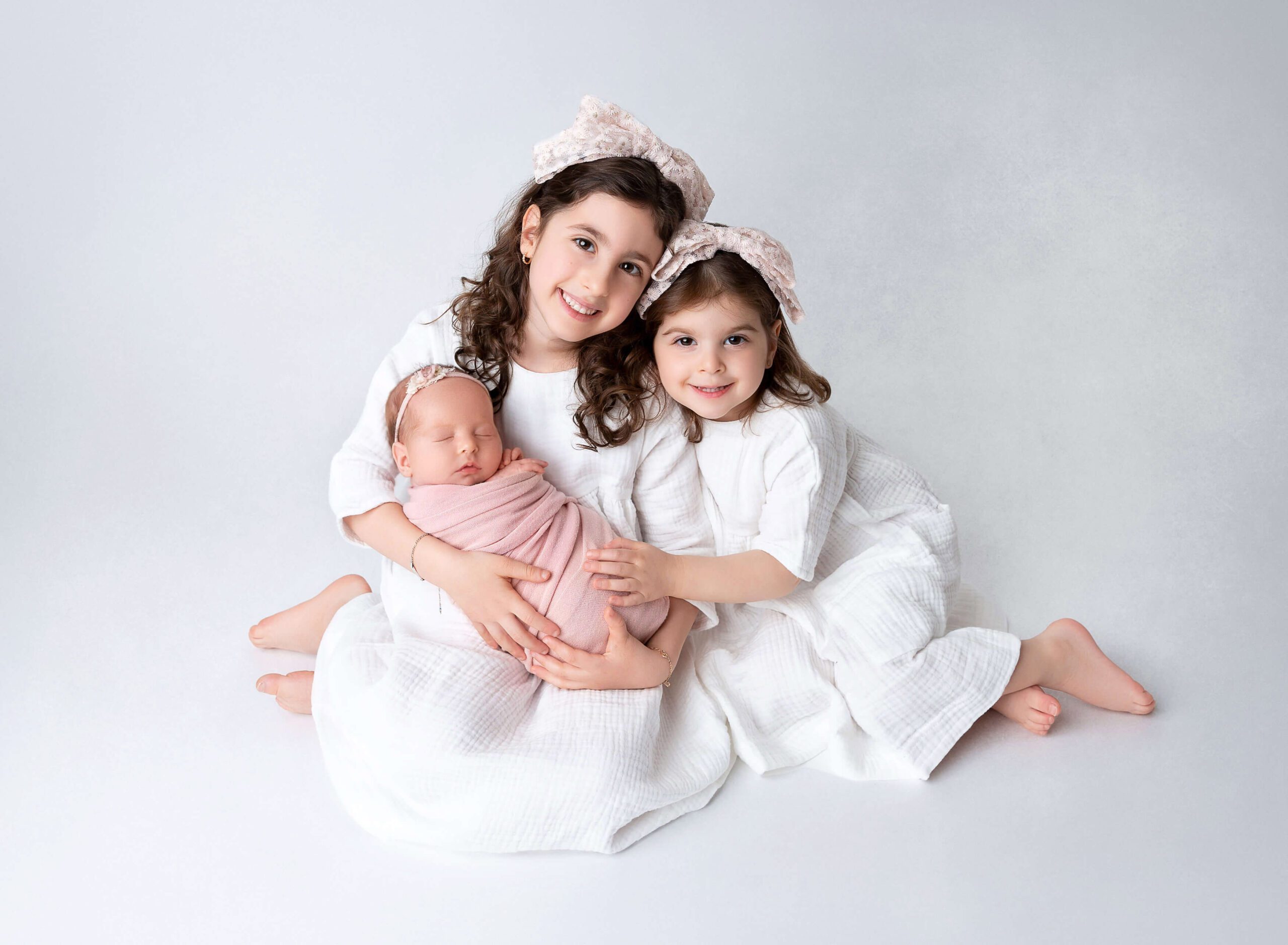 Toddler sisters sit on the floor of a studio holding their sleeping newborn sister in a pink swaddle after using Everything But The Baby in Brooklyn